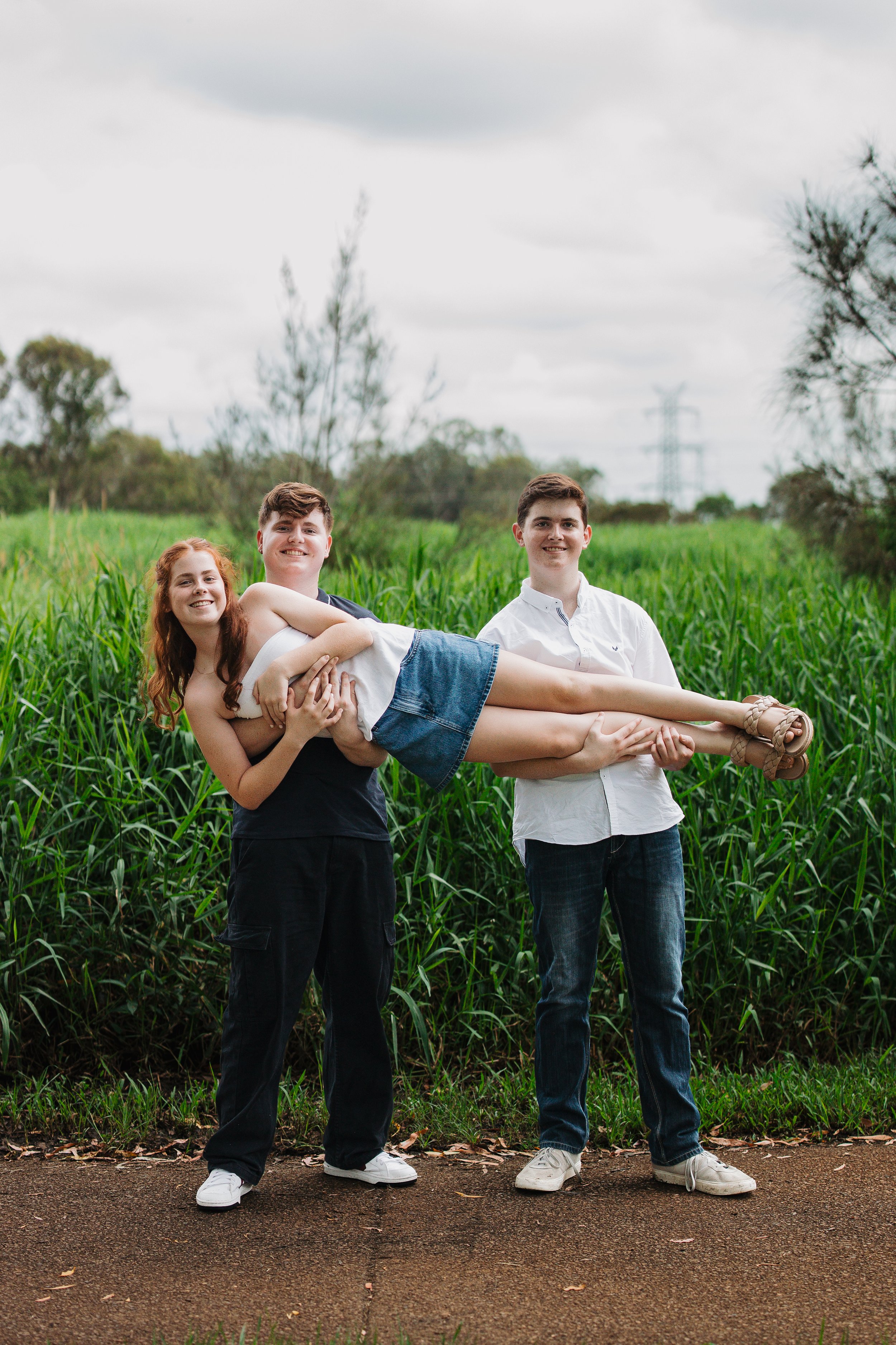 Three young people standing outdoors with lush green grass and trees in the background, one girl being supported horizontally by two boys, all smiling.