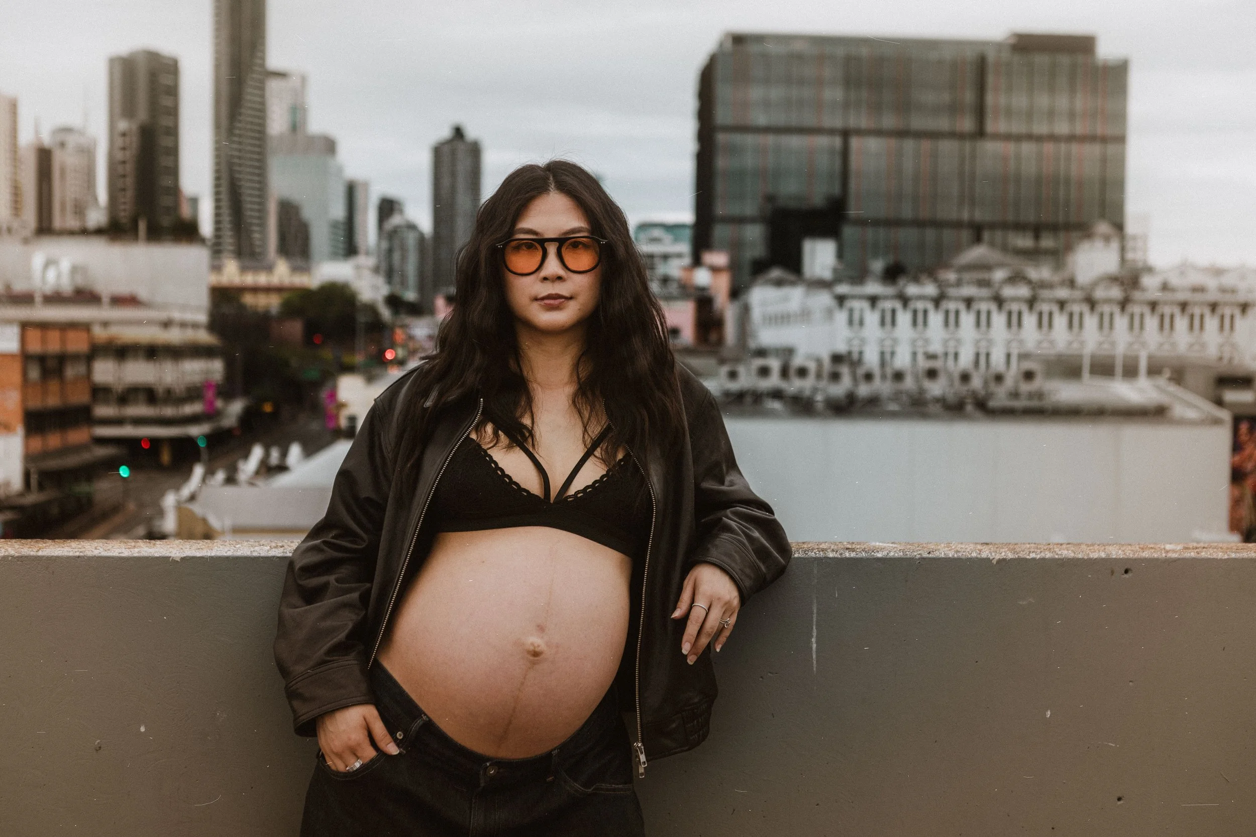 A pregnant woman standing on a rooftop with city buildings in the background, wearing sunglasses, a black jacket, and a black bra, with her hand resting on her stomach.