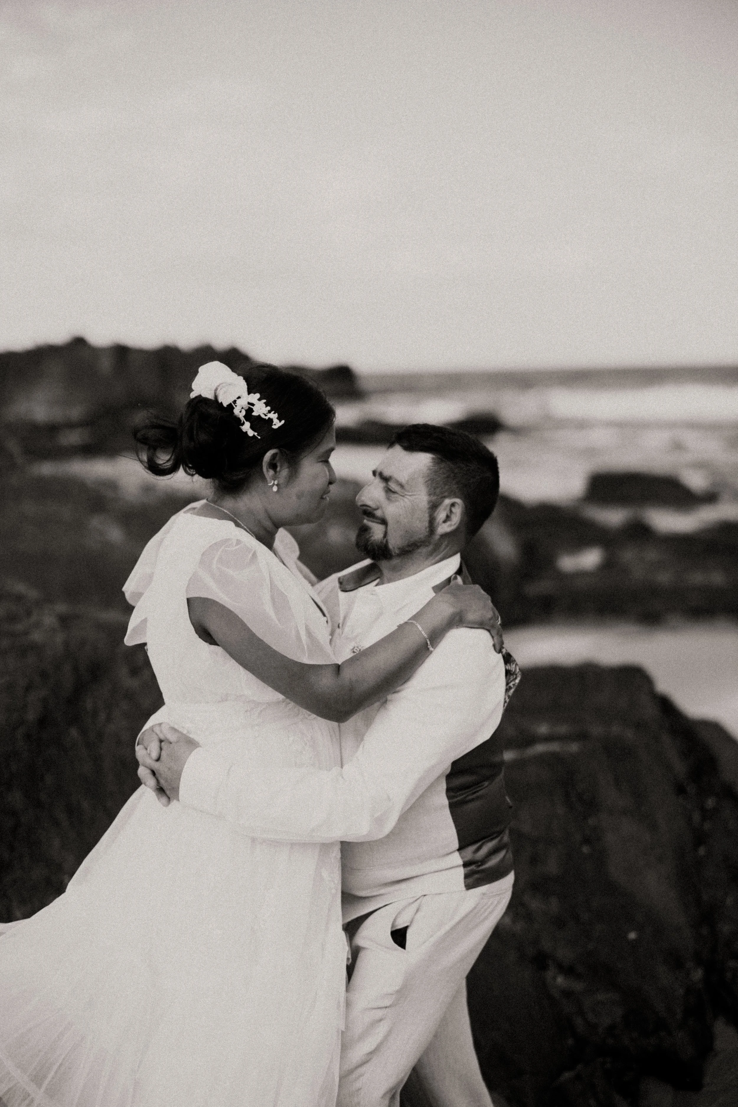 A black-and-white photo of a couple embracing on the beach, with the woman in a white dress and the man in a white shirt and vest, rocky shoreline in the background.
