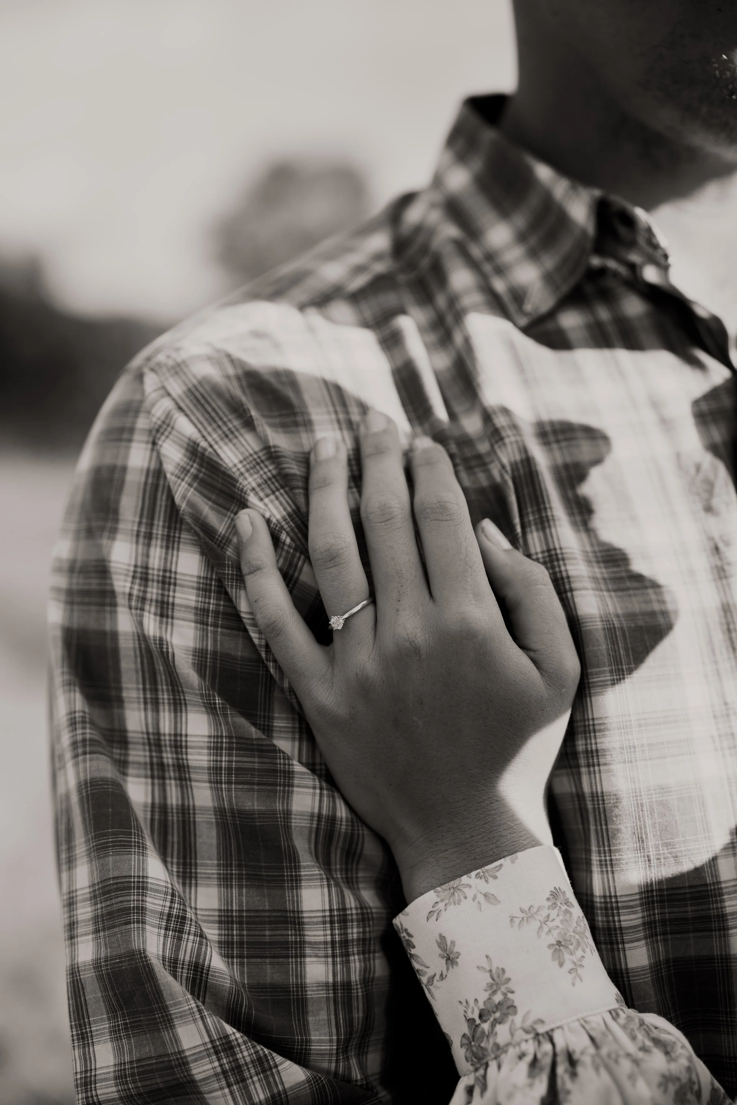 A woman's hand with a ring on her finger is placed on a man's shoulder, who is wearing a plaid shirt. The photo is in black and white.