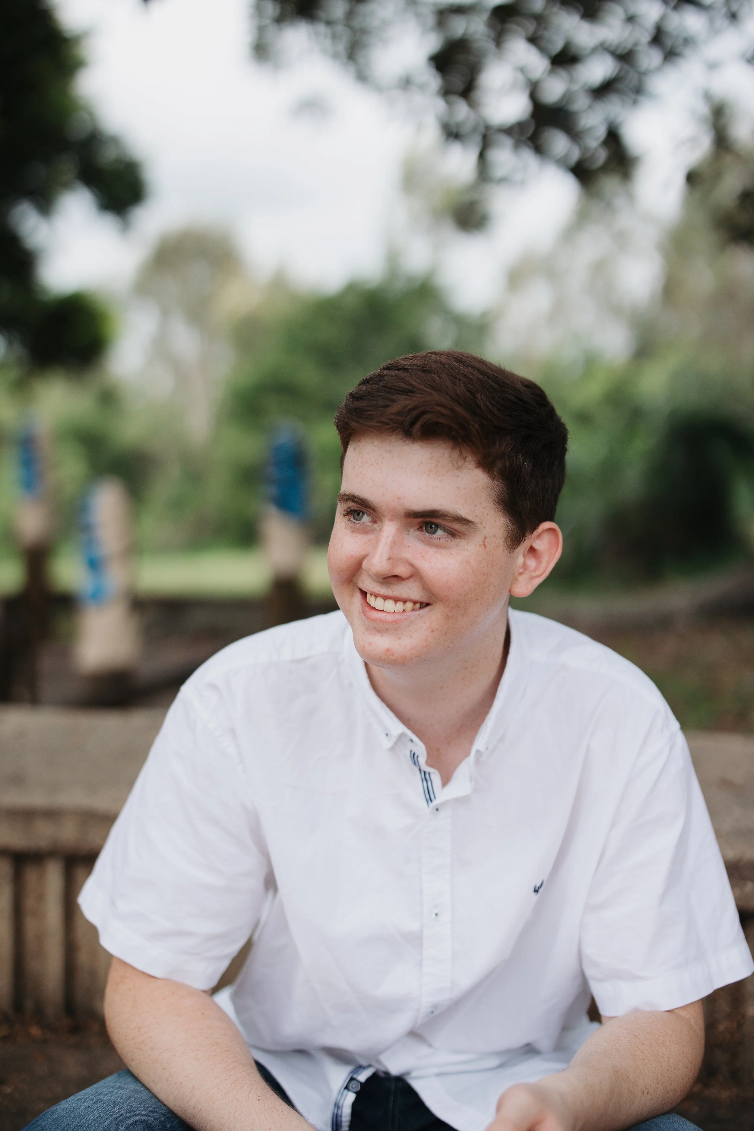 A young man with short brown hair and freckles, wearing a white shirt, sitting outdoors on a wooden bench, smiling and looking to his right, with trees and blue decorations in the background.