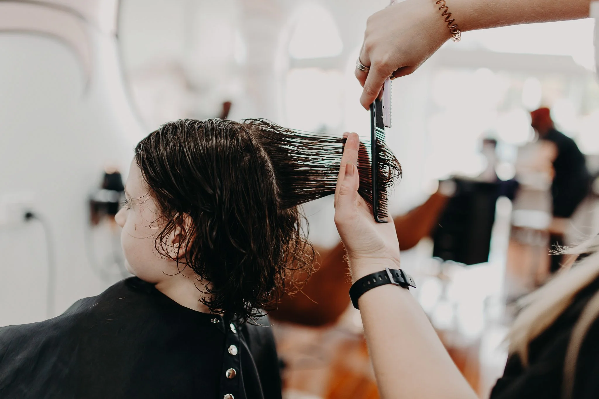 A woman getting her hair styled in a salon, with a hairstylist using a comb and scissors to cut wet, dark hair.