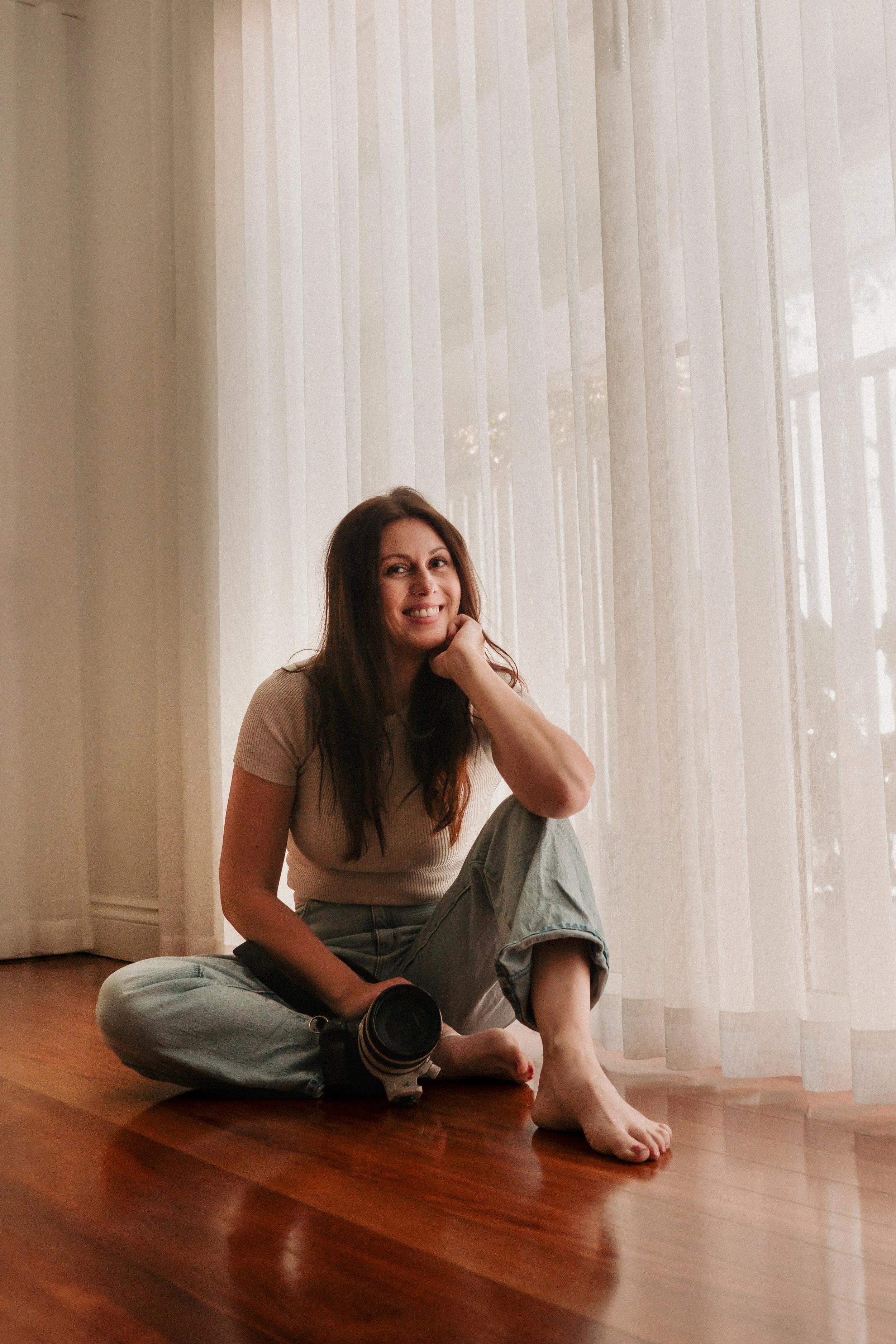 A woman sitting on wooden floor near sheer curtains, smiling at the camera, with a camera resting on the floor beside her.