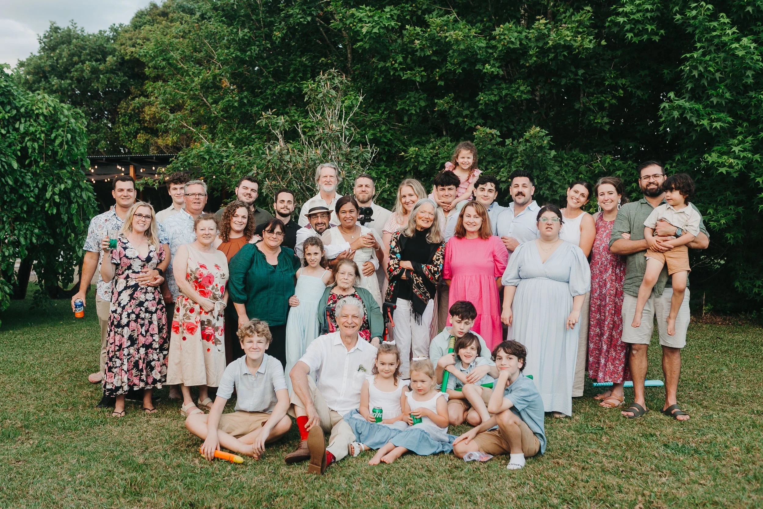 Group of diverse family members and friends gathered outdoors on a grassy lawn for a celebration, with trees and string lights in the background.