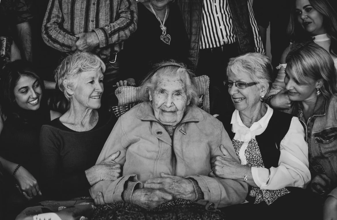 A group of women, including an elderly woman in a wheelchair, smiling and gathered around her in a celebration or family gathering.