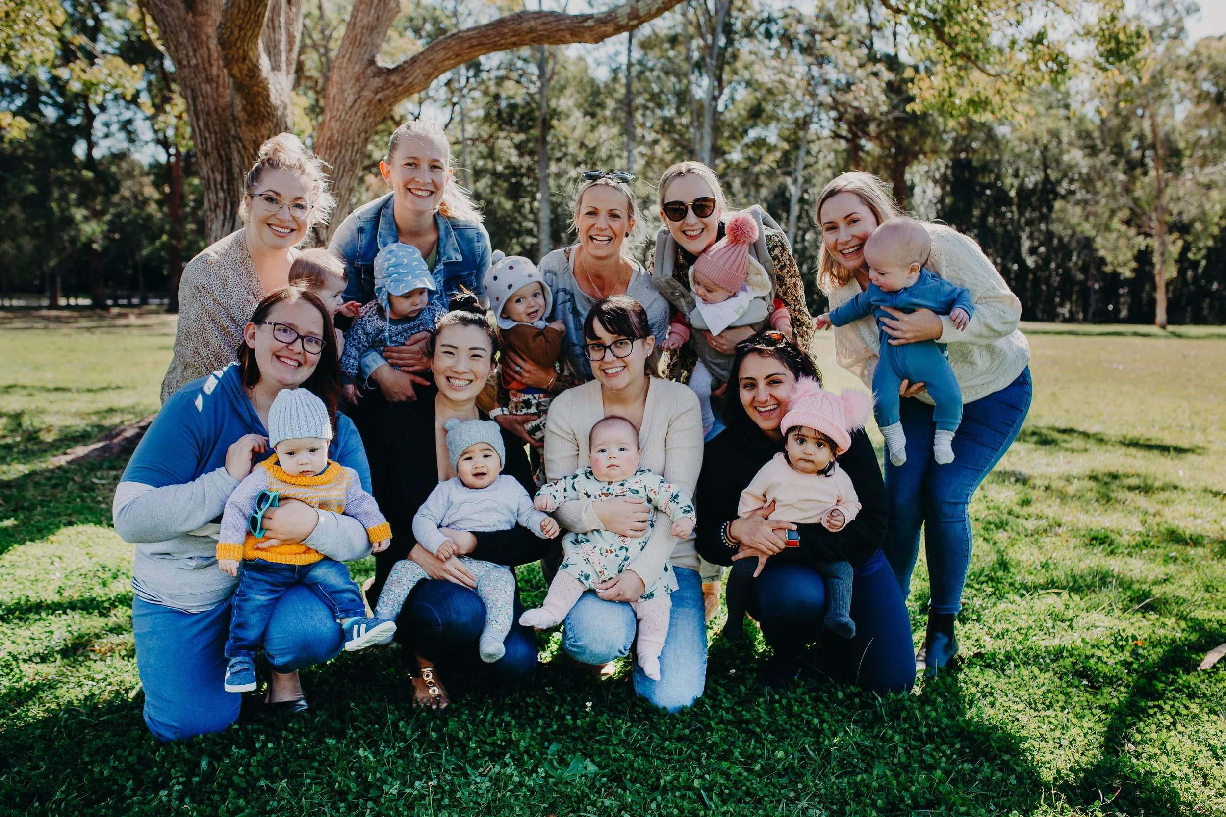 A group of women and children smiling and posing outdoors on a sunny day with trees in the background.