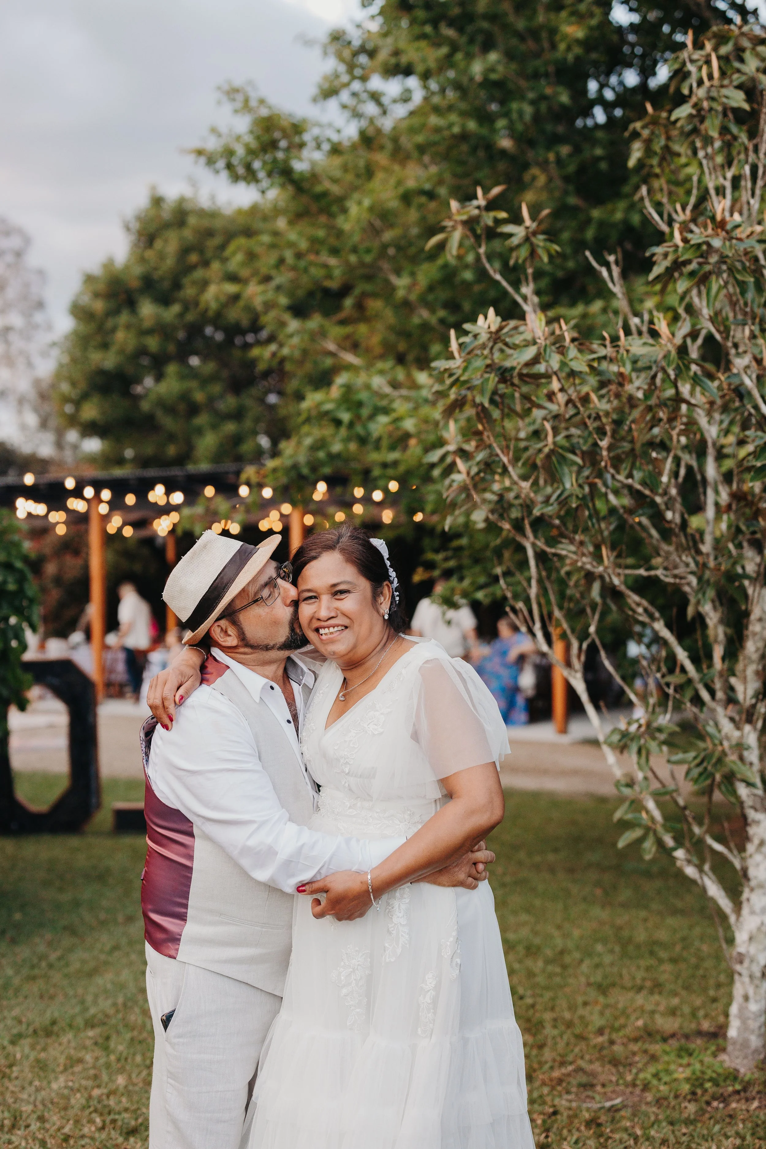 A man in a hat and glasses is kissing a woman in a white dress on the cheek at an outdoor event during the evening. The woman is smiling, and they are embracing each other. There are string lights and other people in the background.