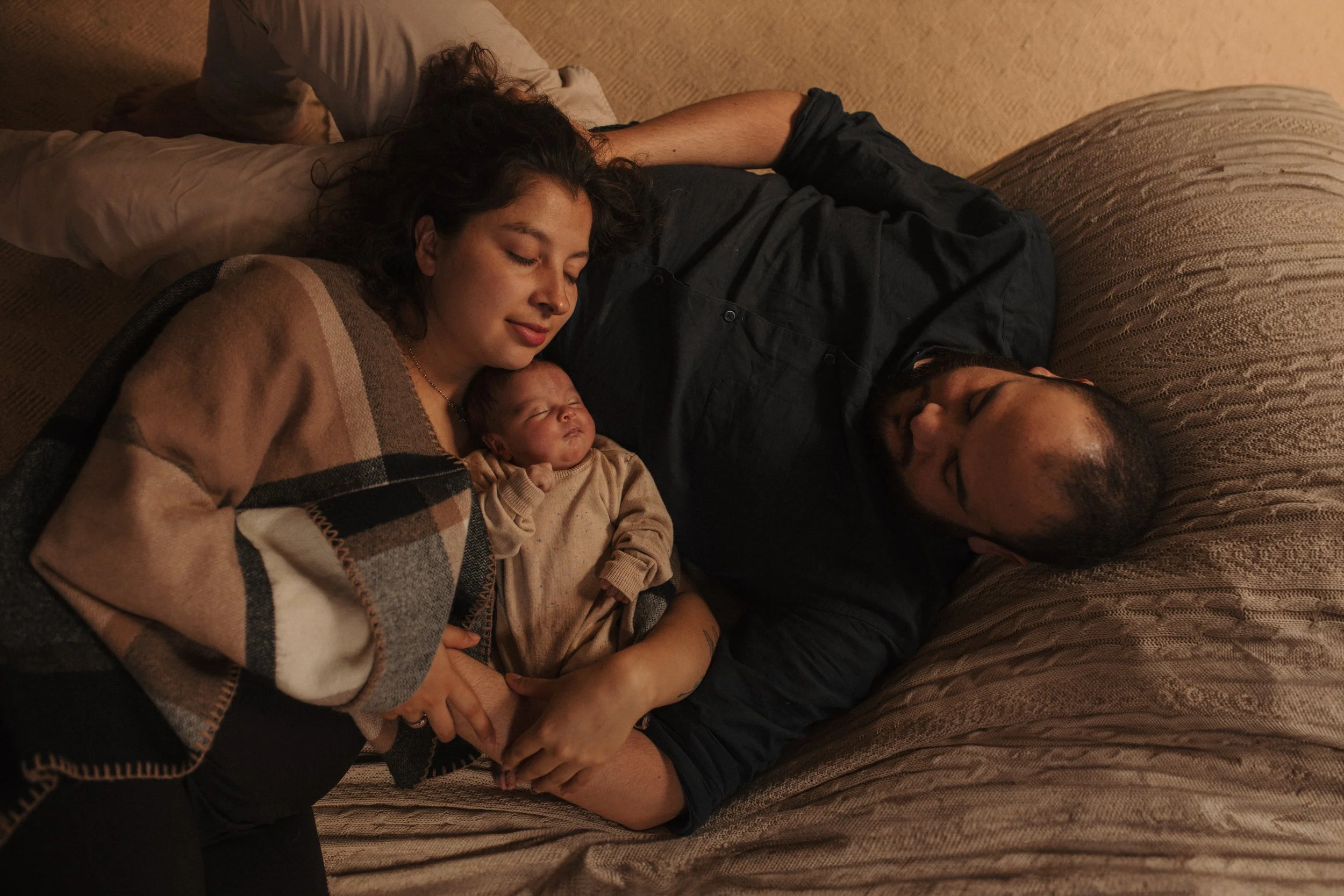 A woman and a man are lying on a couch, sleeping peacefully. The woman is holding a small baby close to her chest, also asleep. The woman is wearing a checkered sweater and the man is dressed in a dark shirt. The scene is warm and cozy, set in a dimly lit room.
