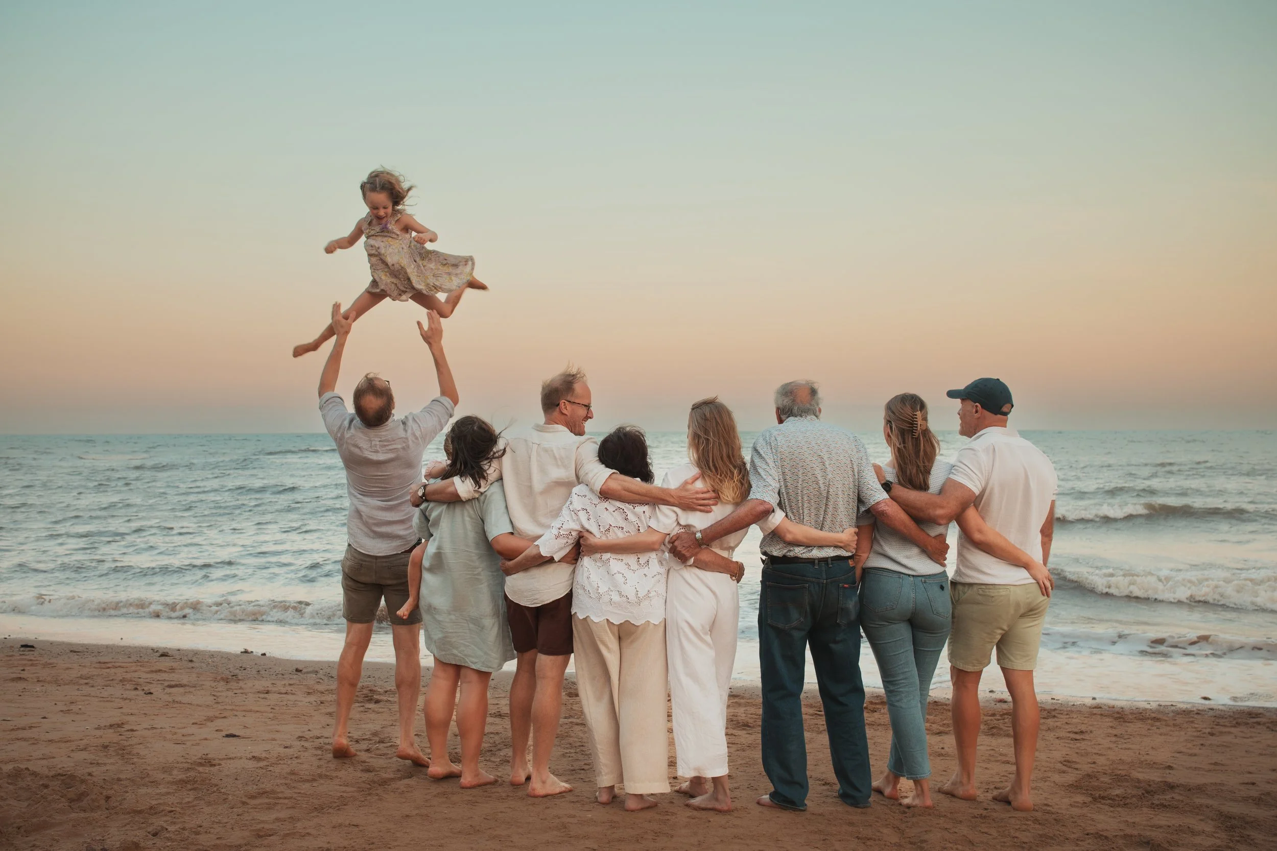 Group of nine people, including children, hugging and interacting on a sandy beach during sunset, with one person lifting a young girl in the air.