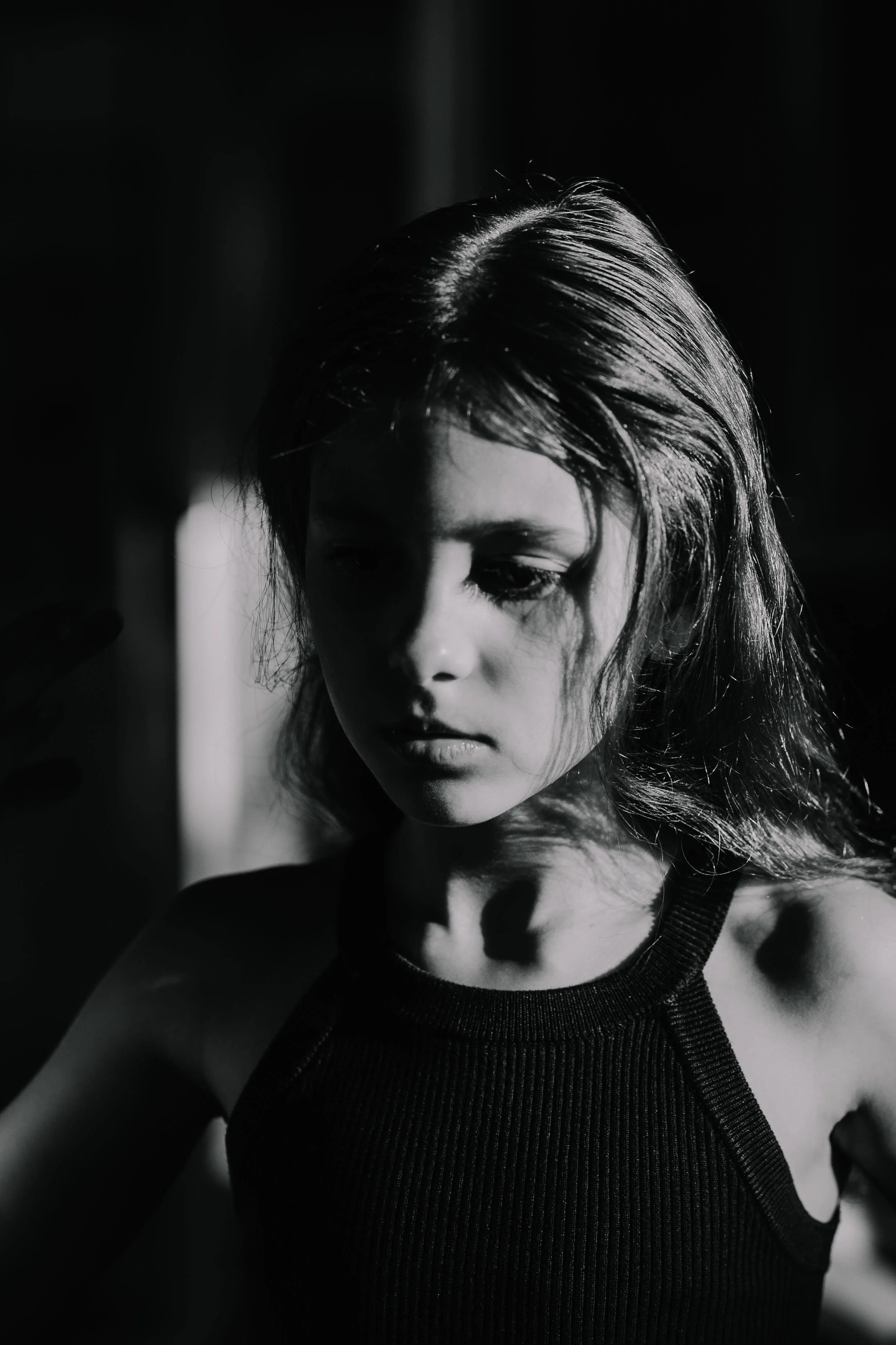 Black and white photo of a young girl with wet hair, wearing a sleeveless top, looking down thoughtfully with shadows on her face.