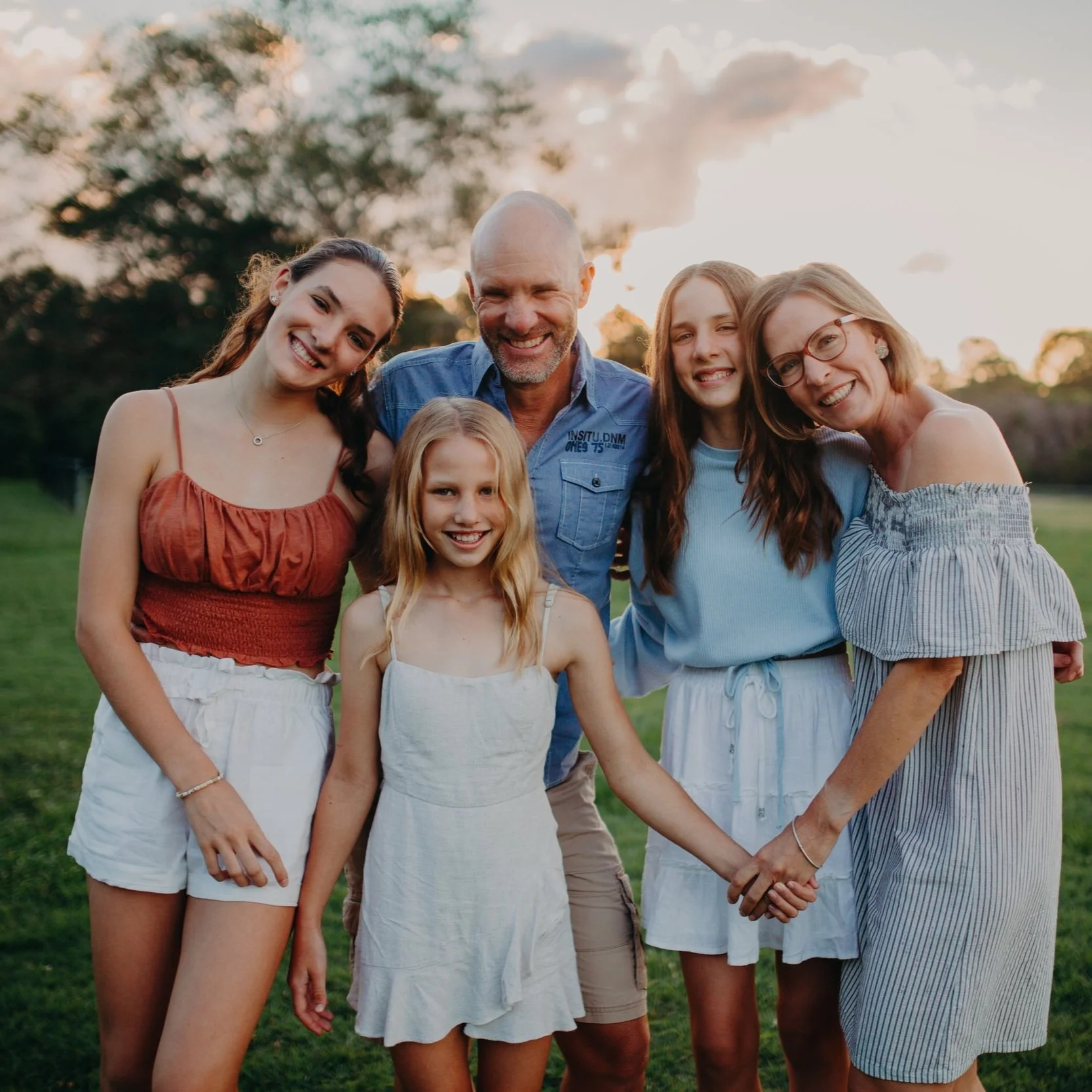 Six family members, including children and adults, smiling and holding hands outdoors during sunset.