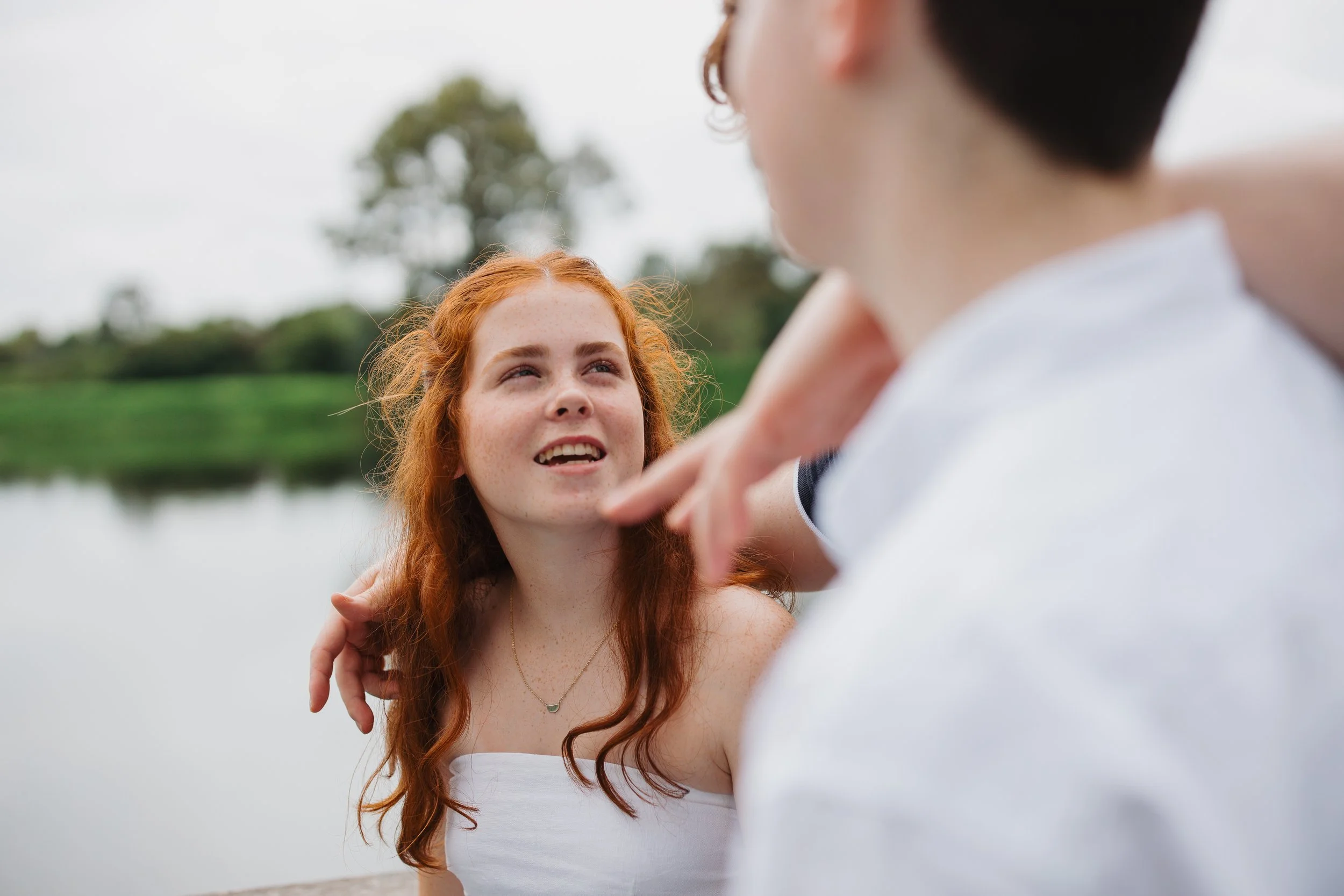 A red-haired woman smiling at a man outdoors near a body of water with green trees in the background.