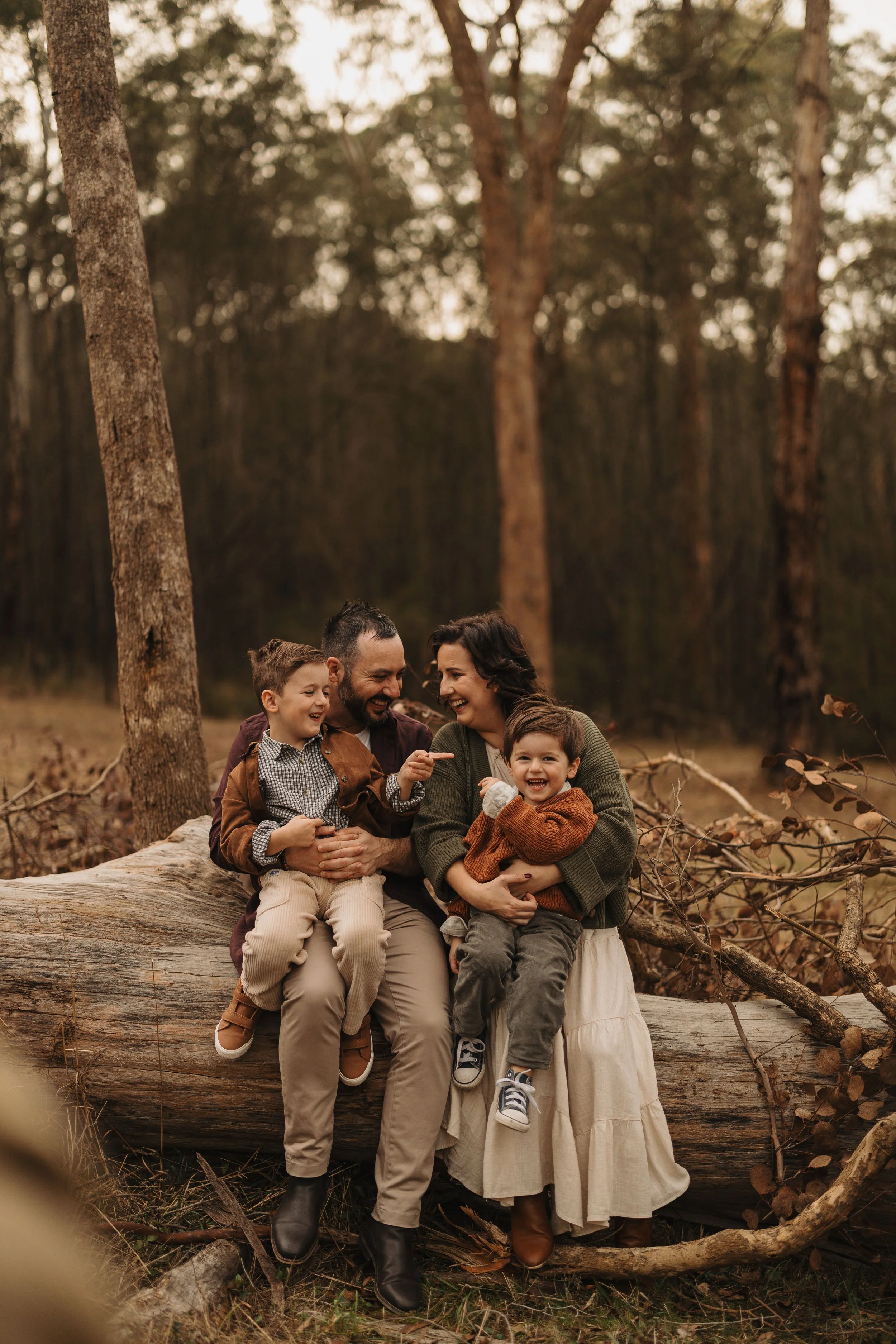 A family of four, including two children, sits on a fallen log in a forest, smiling and enjoying each other's company.