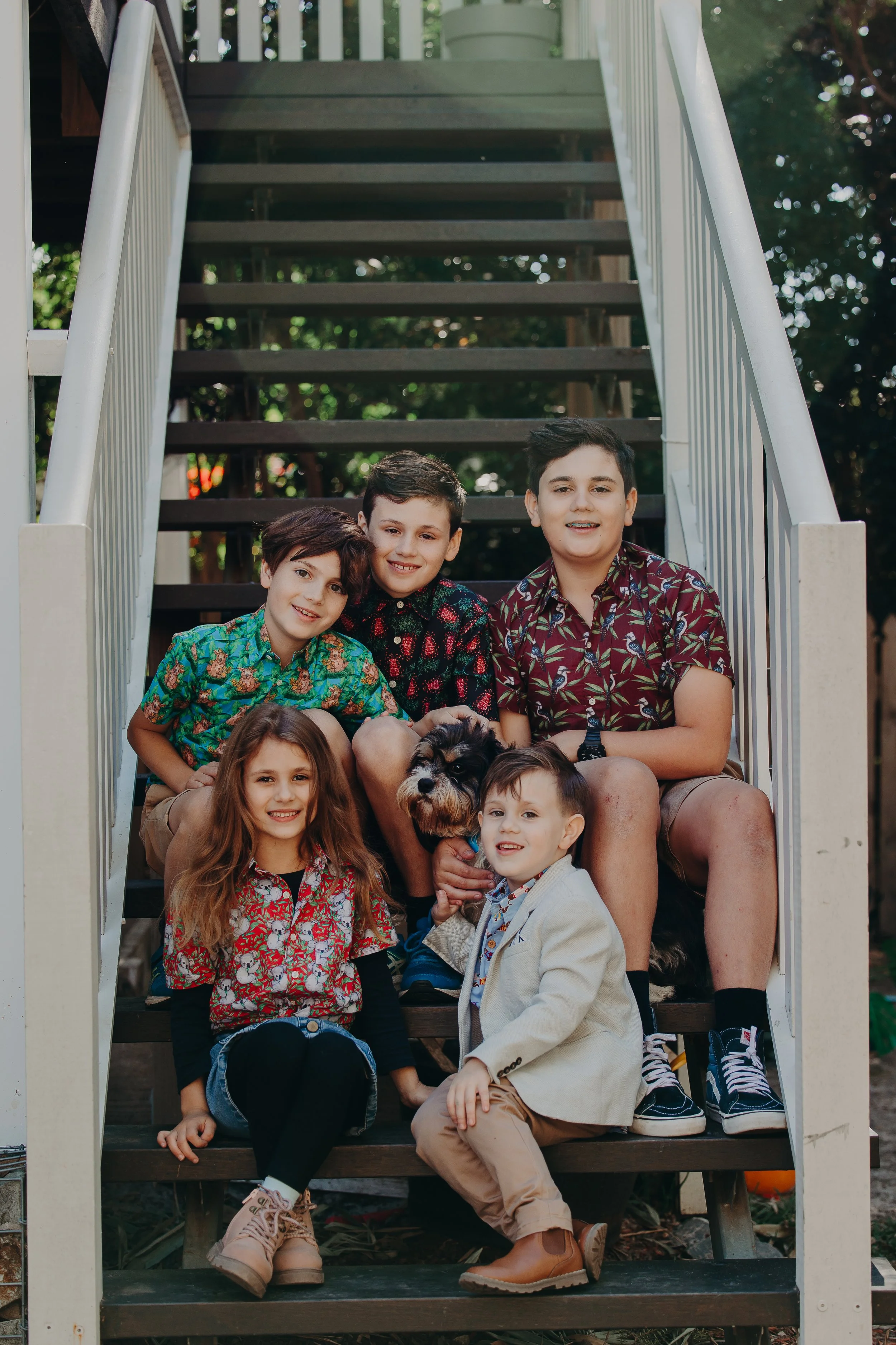 A group of six children and a dog sitting on outdoor staircase with trees in background.