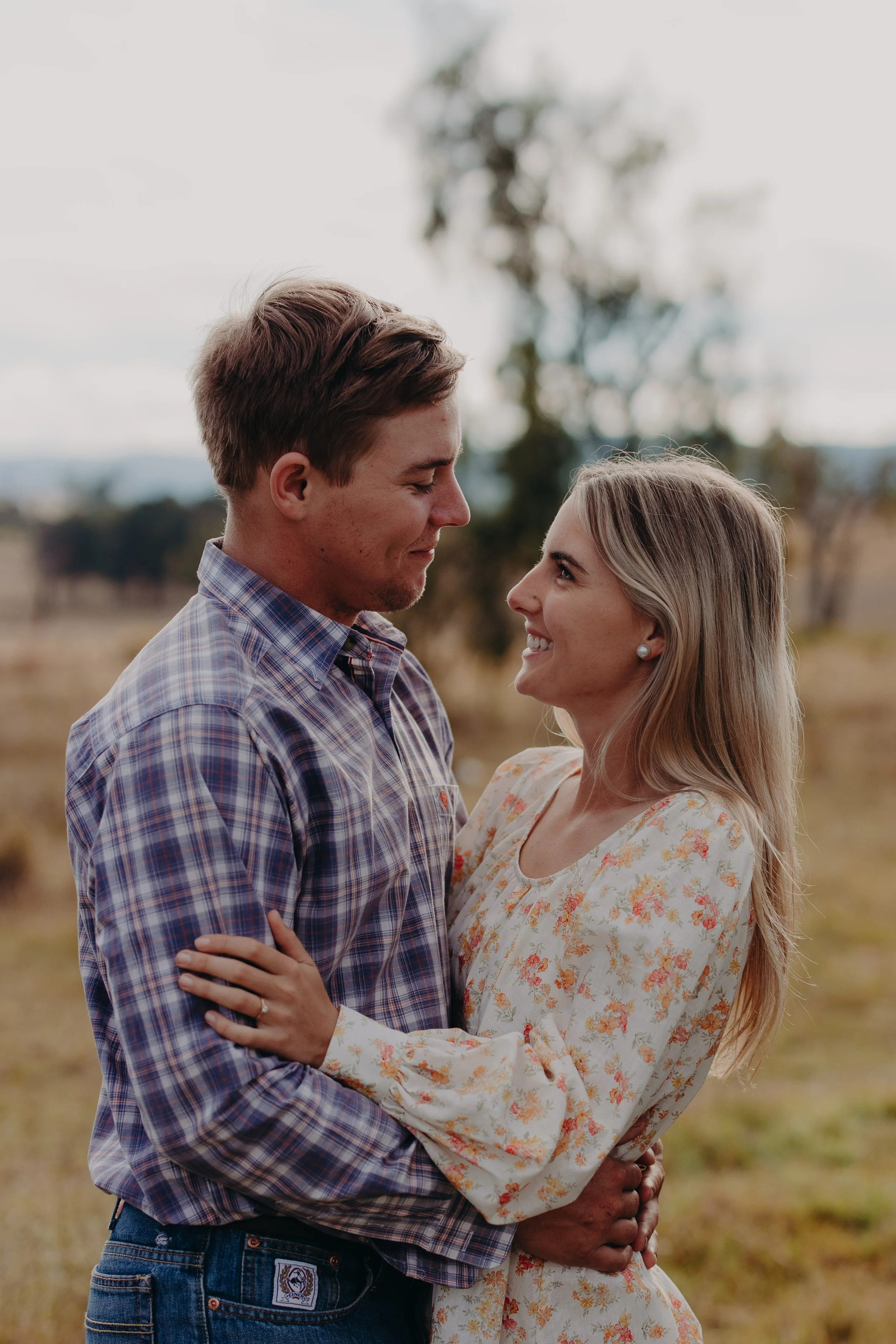 A young couple standing close together outdoors, smiling and embracing each other in a grassy field with trees in the background.