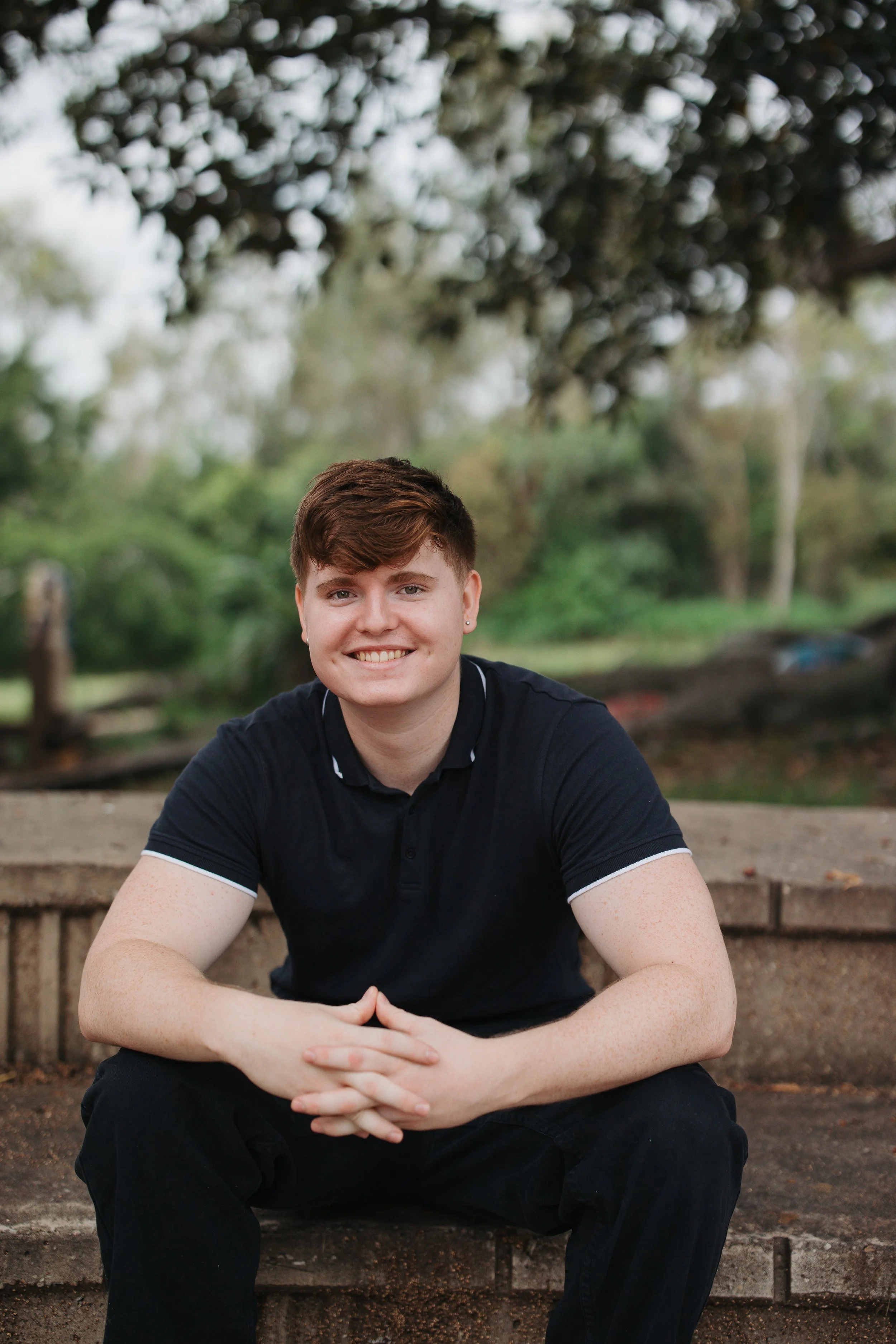 A young man with short brown hair and light skin, wearing a black polo shirt, sitting outdoors on a brick ledge, smiling with hands clasped, with a blurred green background of trees and foliage.