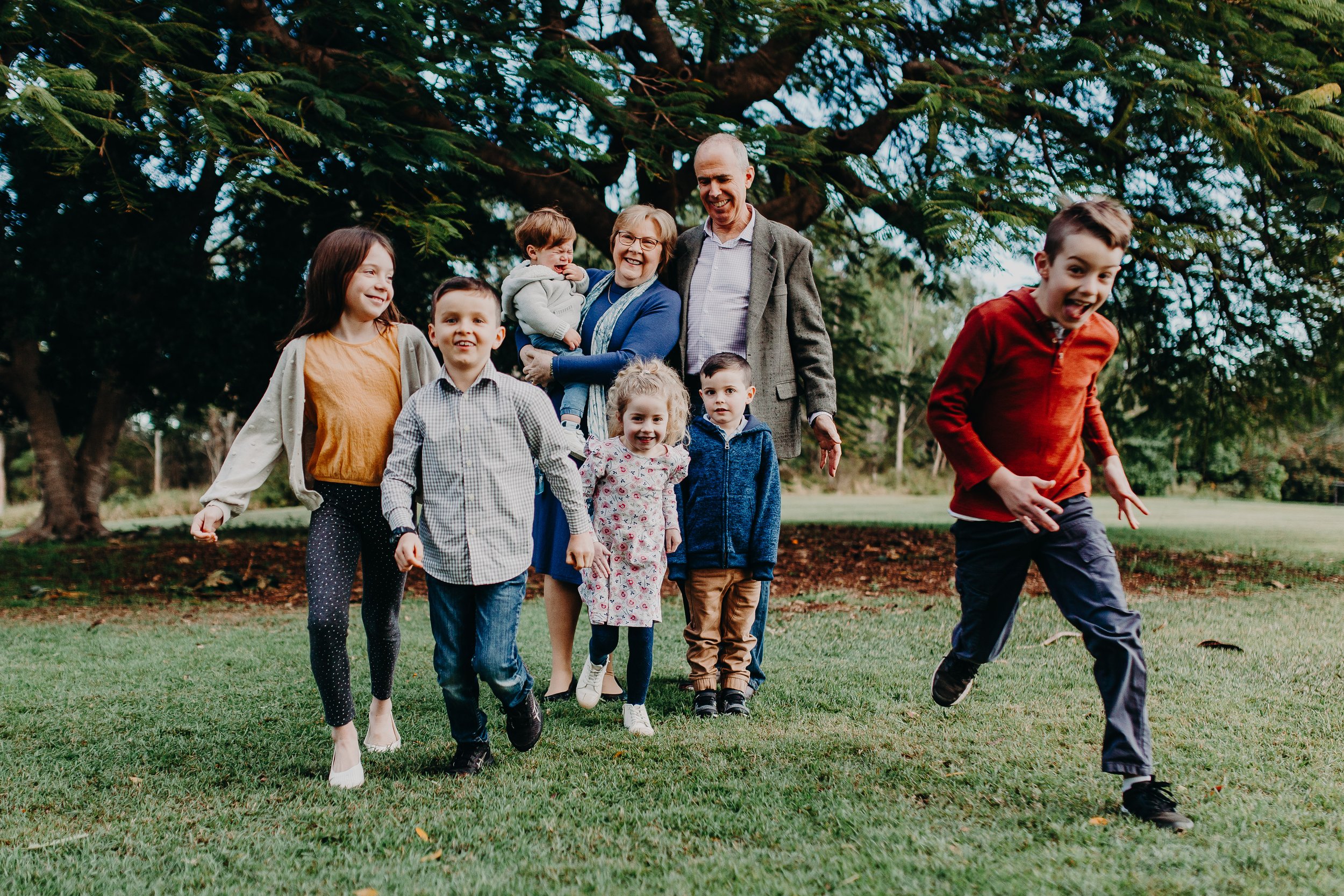 A family of eight, including children and grandparents, is playing and running on a grassy field outdoors near large trees on a clear day.