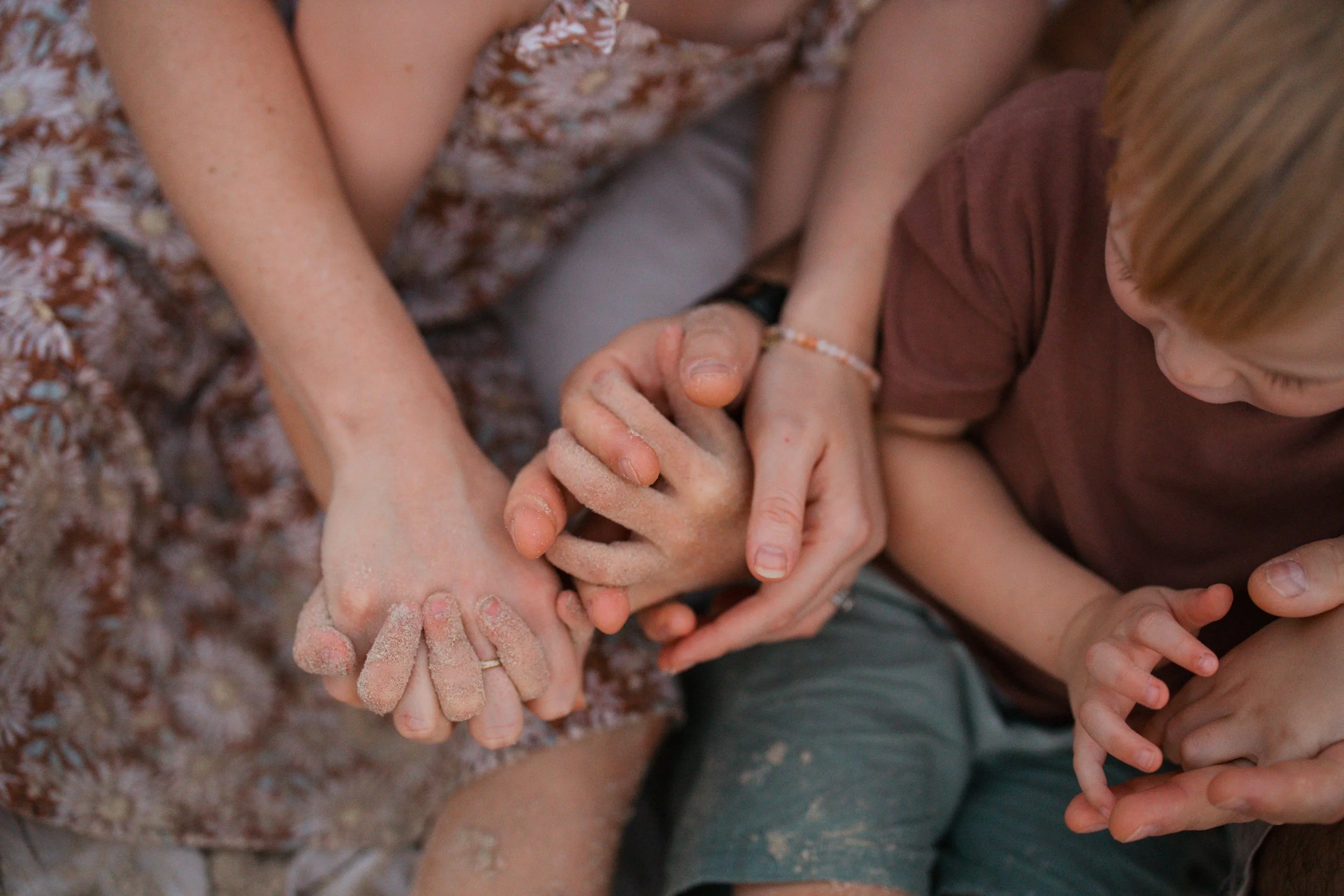 Multiple hands, some with rings and some with dirt, are clasped together in a group hug. A child with reddish hair is visible to the right, looking down at the hands.