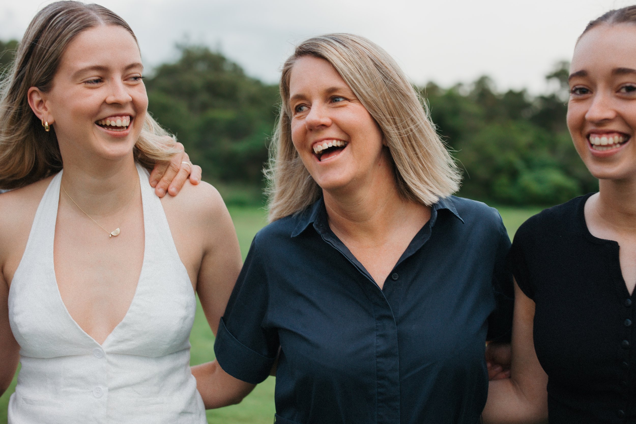 Three women smiling and standing outdoors in a field, with trees and cloudy sky in the background.