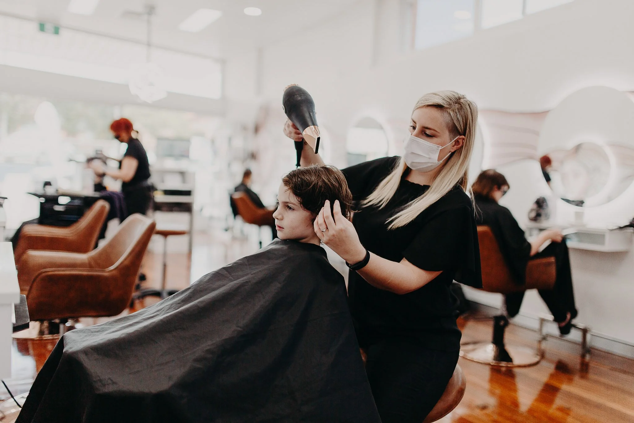 A hairstylist blow-drying a young boy's hair in a modern salon. The stylist and boy are wearing masks. Other clients are visible in the background, getting hair services.