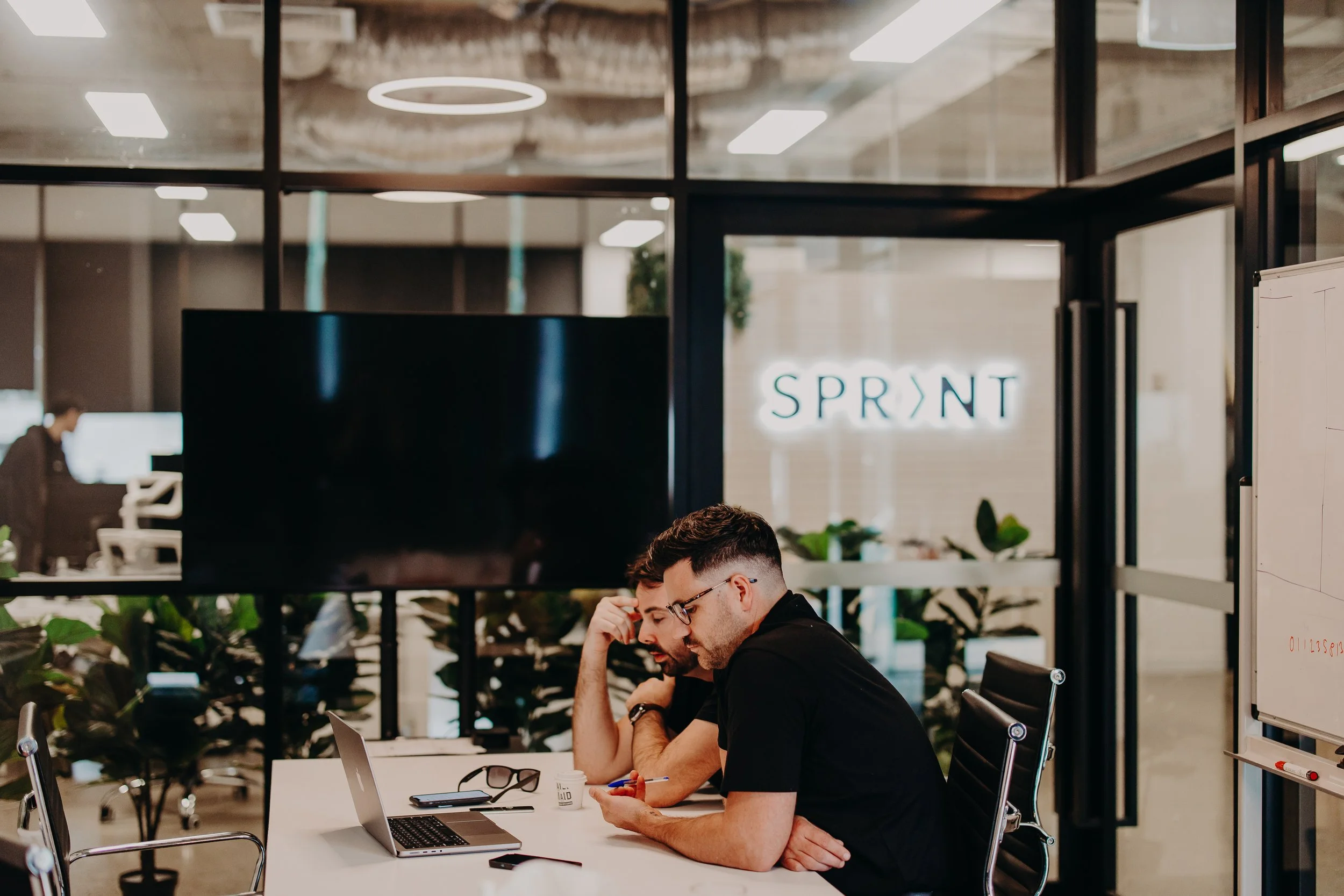 Two men sitting at a white table in a modern office space, working together and looking at a smartphone. On the table are a laptop, glasses, and a mug. Behind them, a glass wall with the word 'SPRINT' illuminated on it, and some plants are visible in