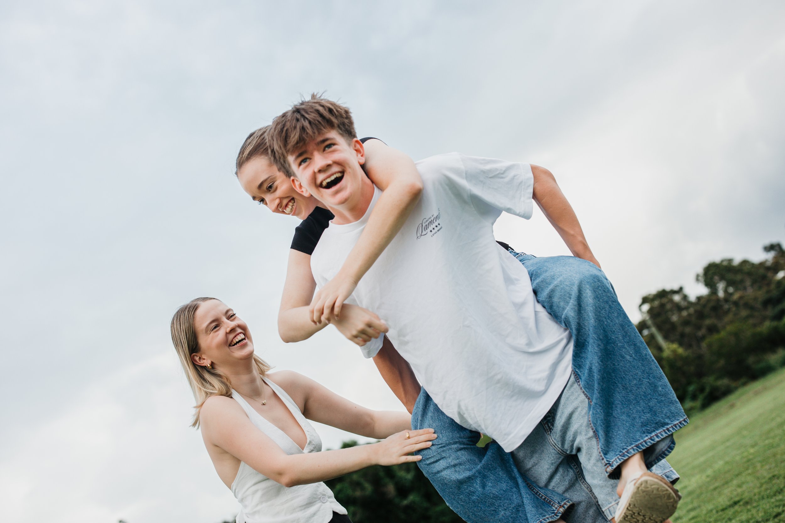 A group of four people, two young boys and two women, playing outdoors on grass under a cloudy sky. One boy is riding on the back of another boy, with the women smiling and reaching out to support them.