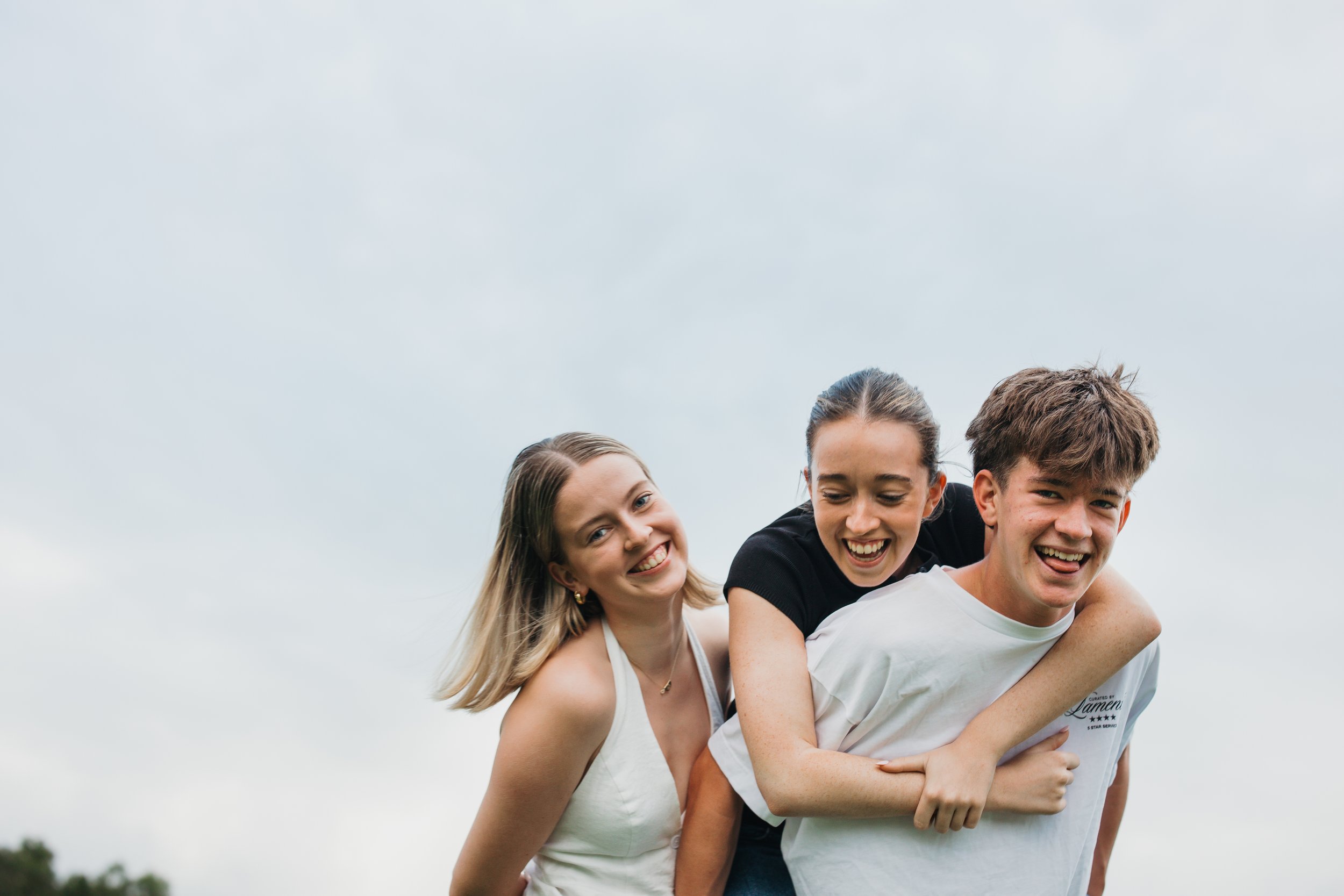 Three young friends smiling and enjoying a piggyback ride outdoors on a cloudy day.