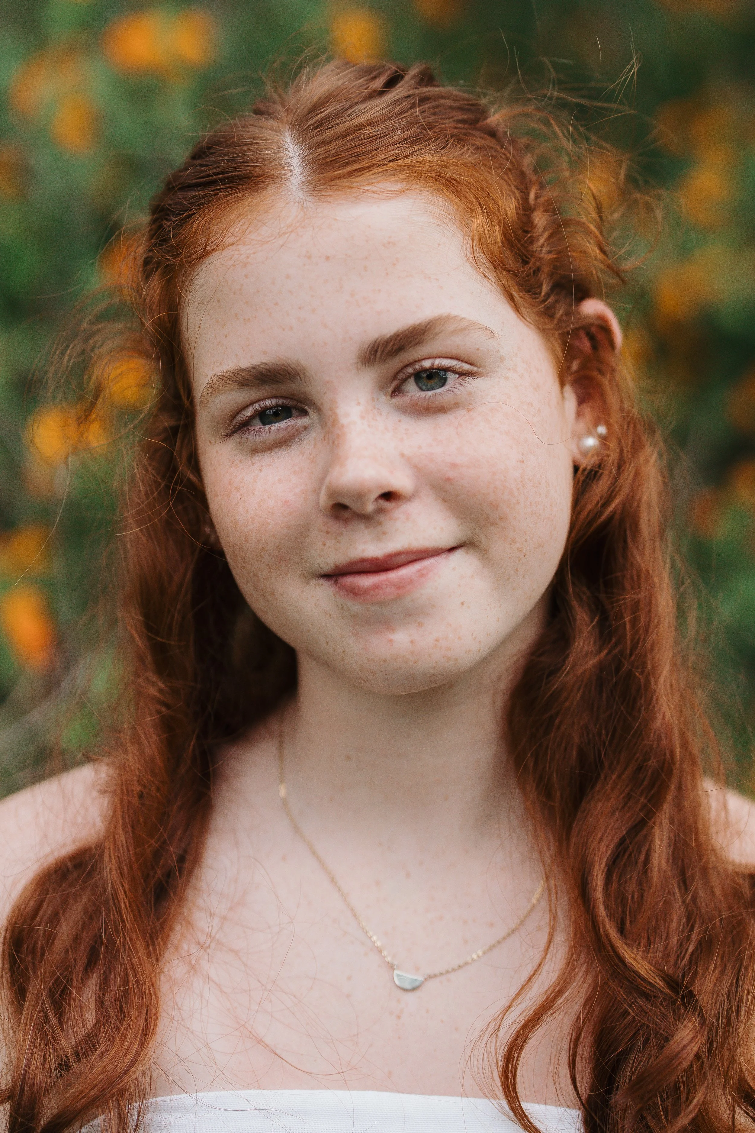 Close-up of a young woman with red hair, freckles, and blue eyes, smiling outdoors with a blurred background of green foliage and yellow flowers.