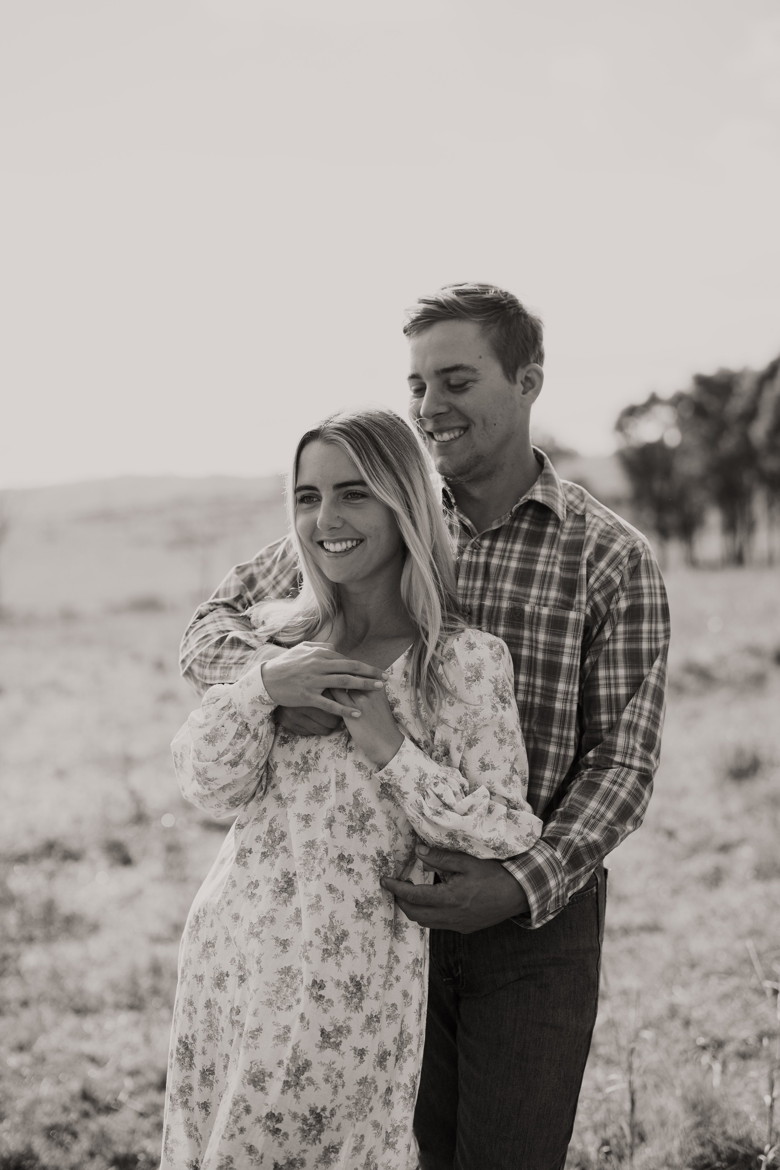 A black-and-white photo of a young couple smiling and embracing outdoors in a field, with trees in the background.