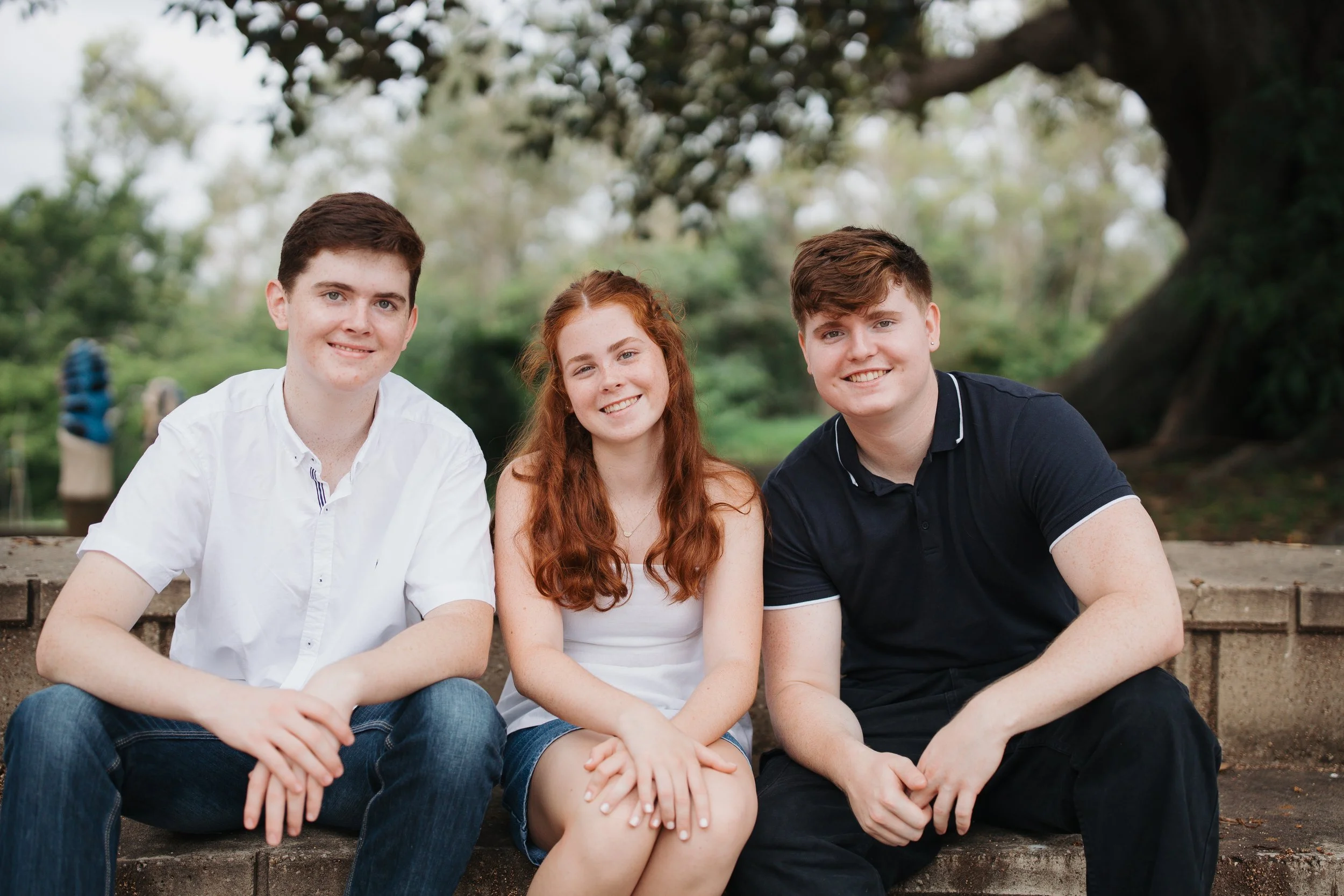 Three teenagers sitting on a concrete ledge outdoors, smiling at the camera. They are in a park with trees in the background.