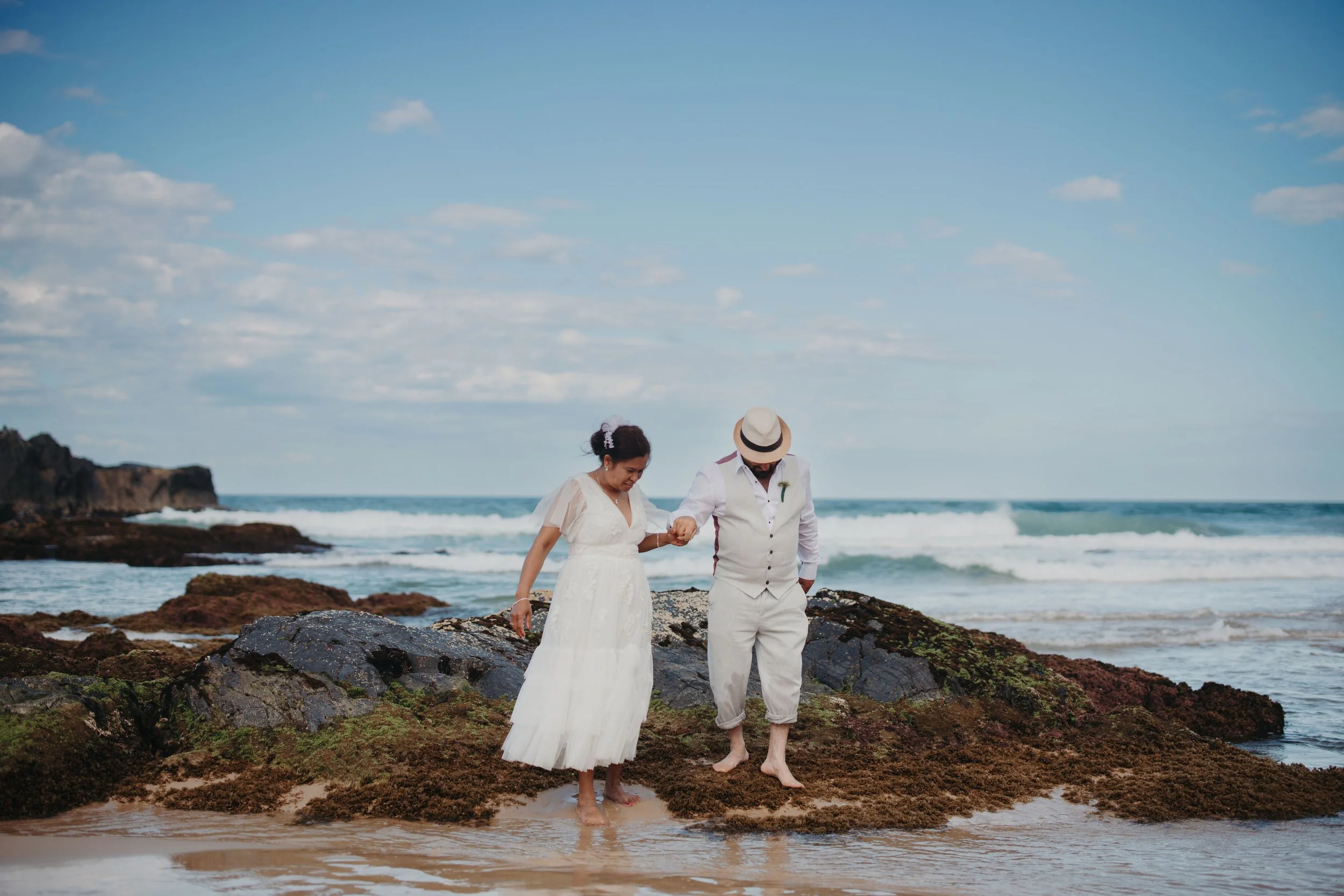 A couple in wedding attire walking barefoot on a rocky beach, holding hands, with the ocean and a partly cloudy sky in the background.