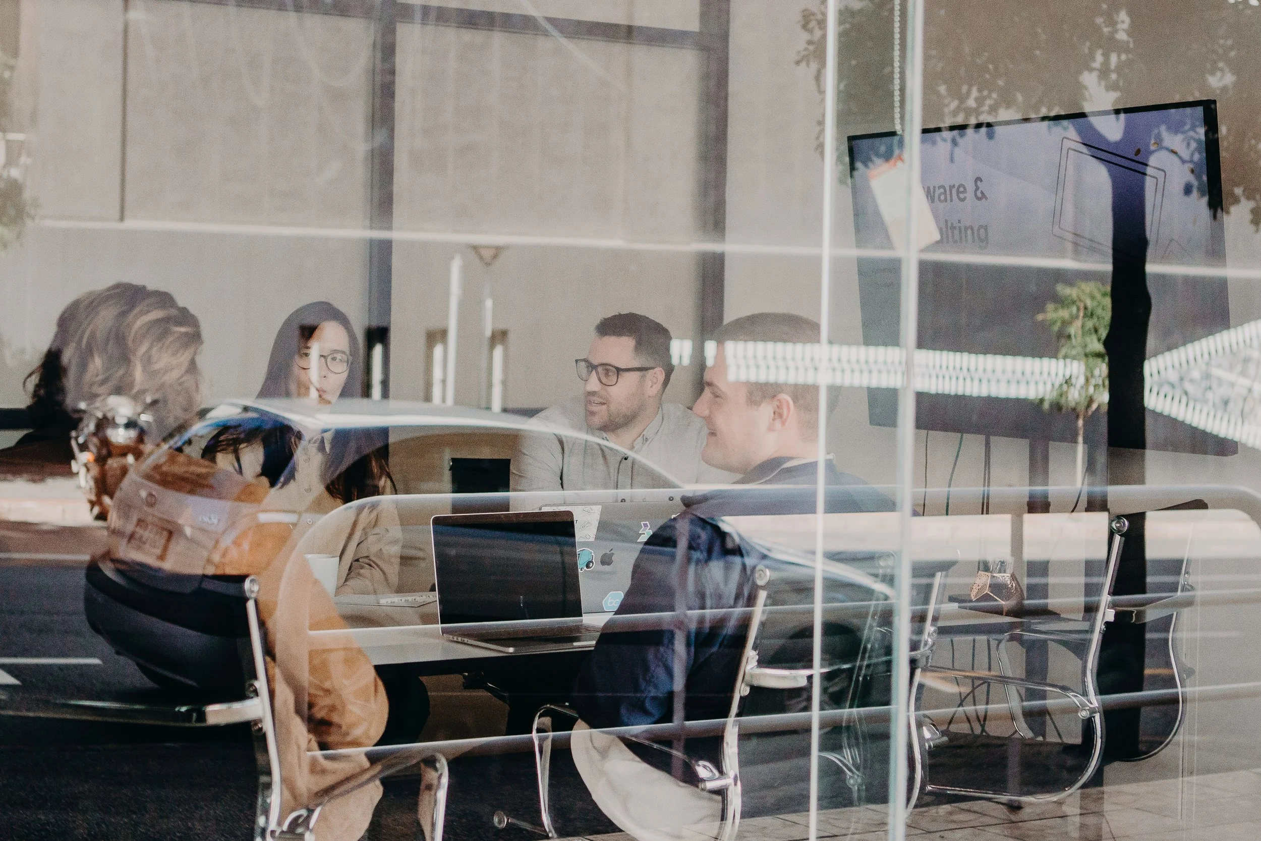 Four people having a business meeting inside an office conference room, with a glass wall showing reflections of the street outside, and a large screen displaying a presentation.