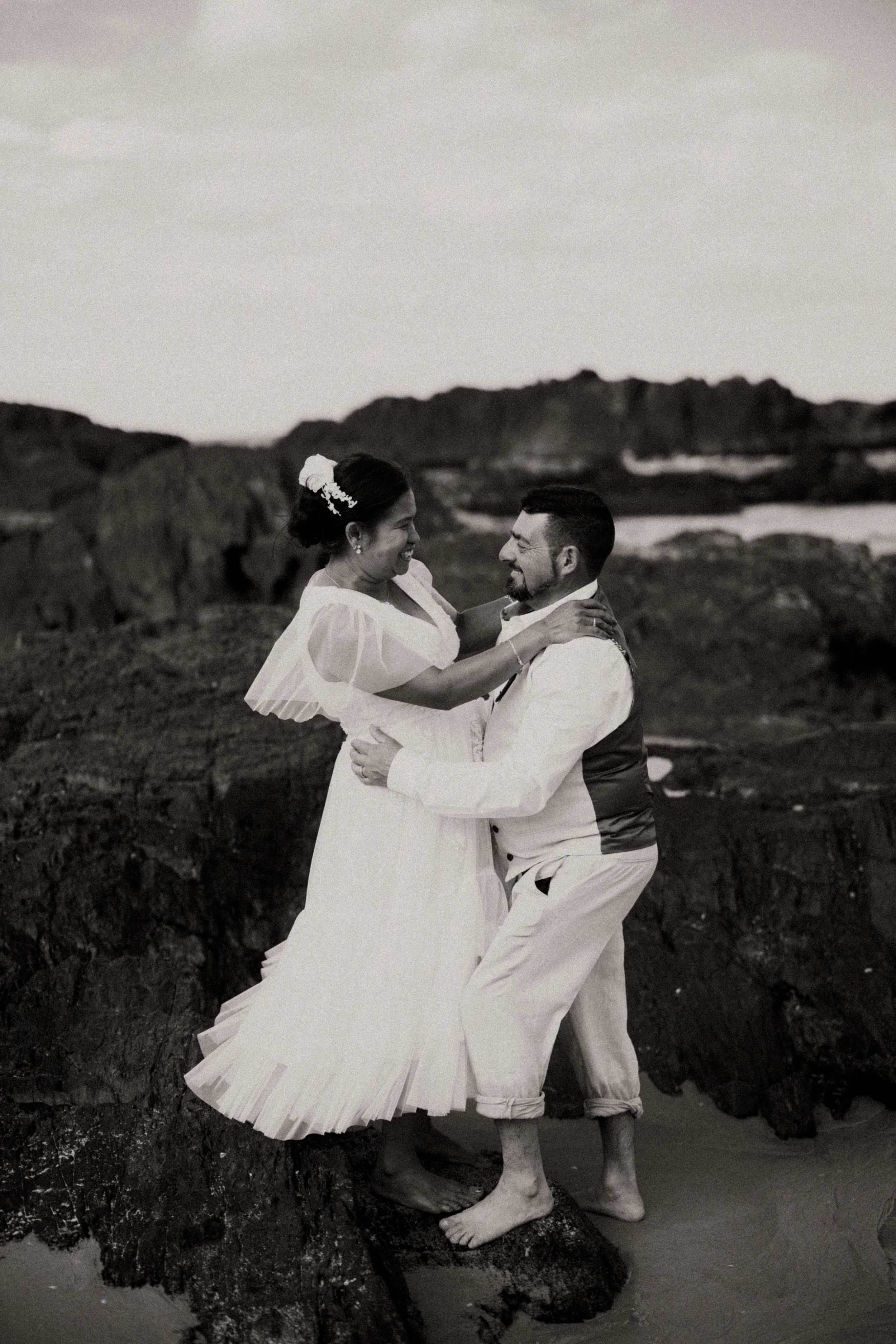 A couple dancing on rocks by the beach, dressed in wedding attire, smiling and enjoying a moment together in black and white.