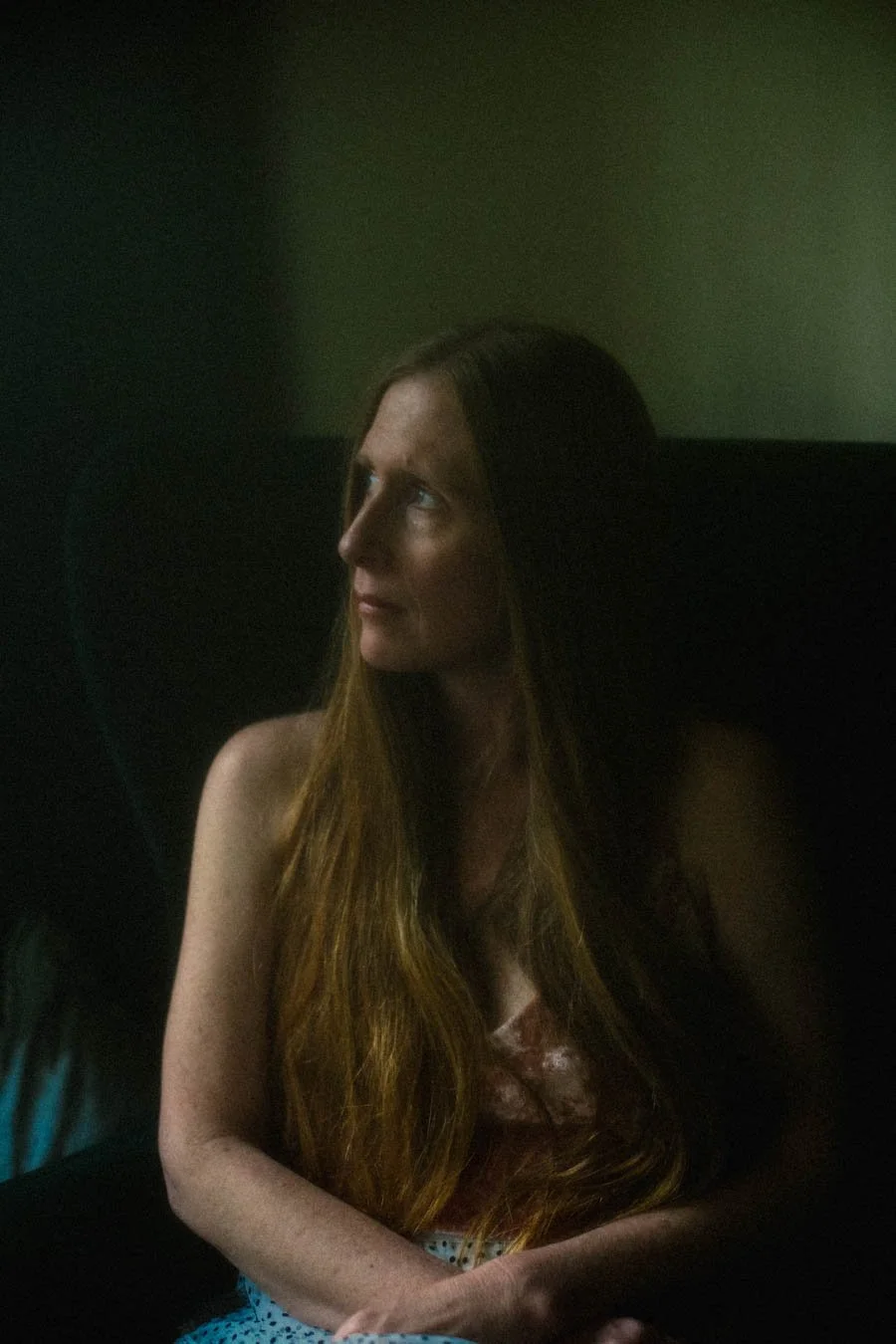 A young woman with long auburn hair sitting in a dark room, looking to the side.