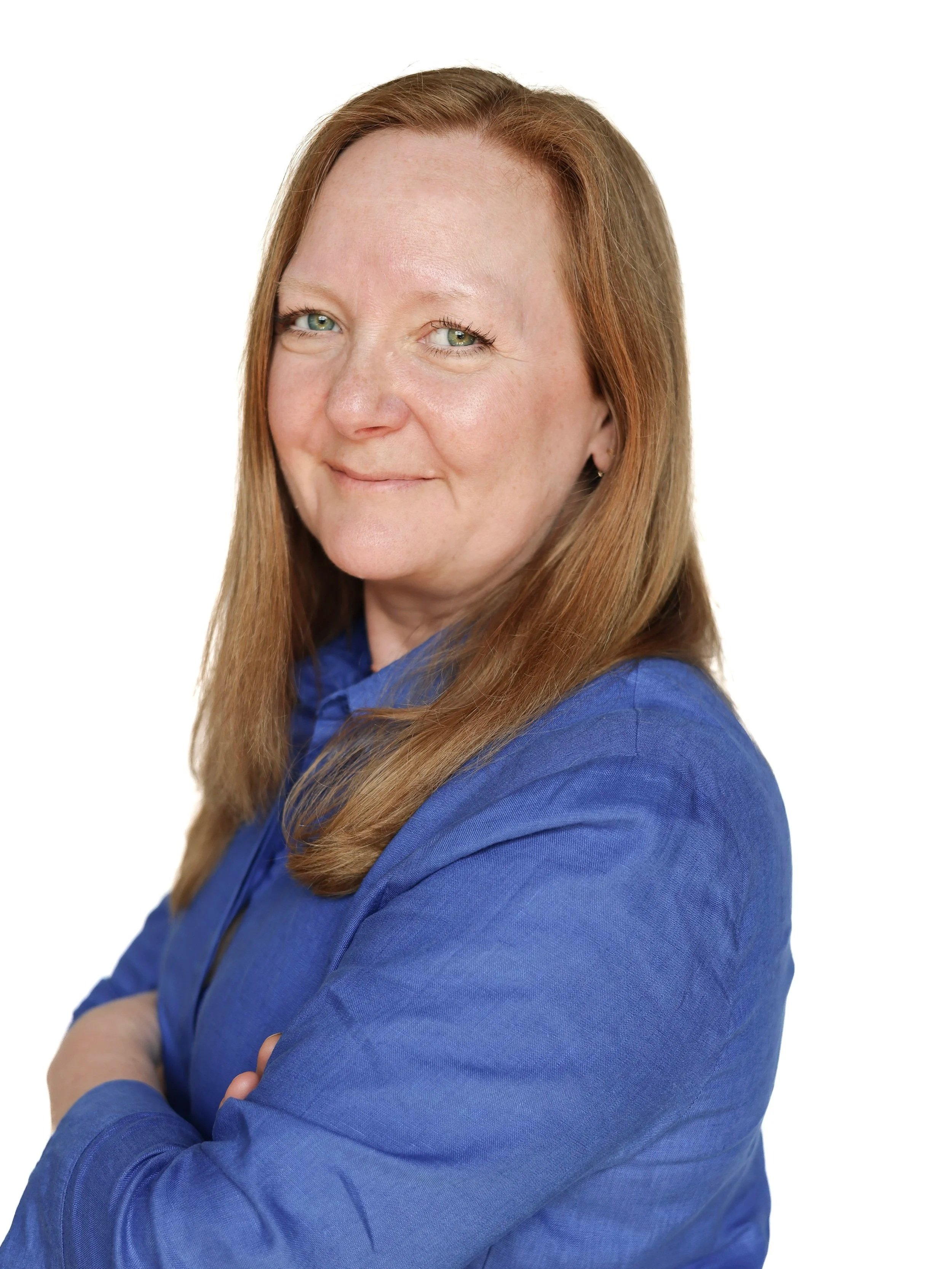 A woman with long red hair, light skin, and green eyes wearing a blue top, standing with arms crossed, smiling slightly, against a plain white background.
