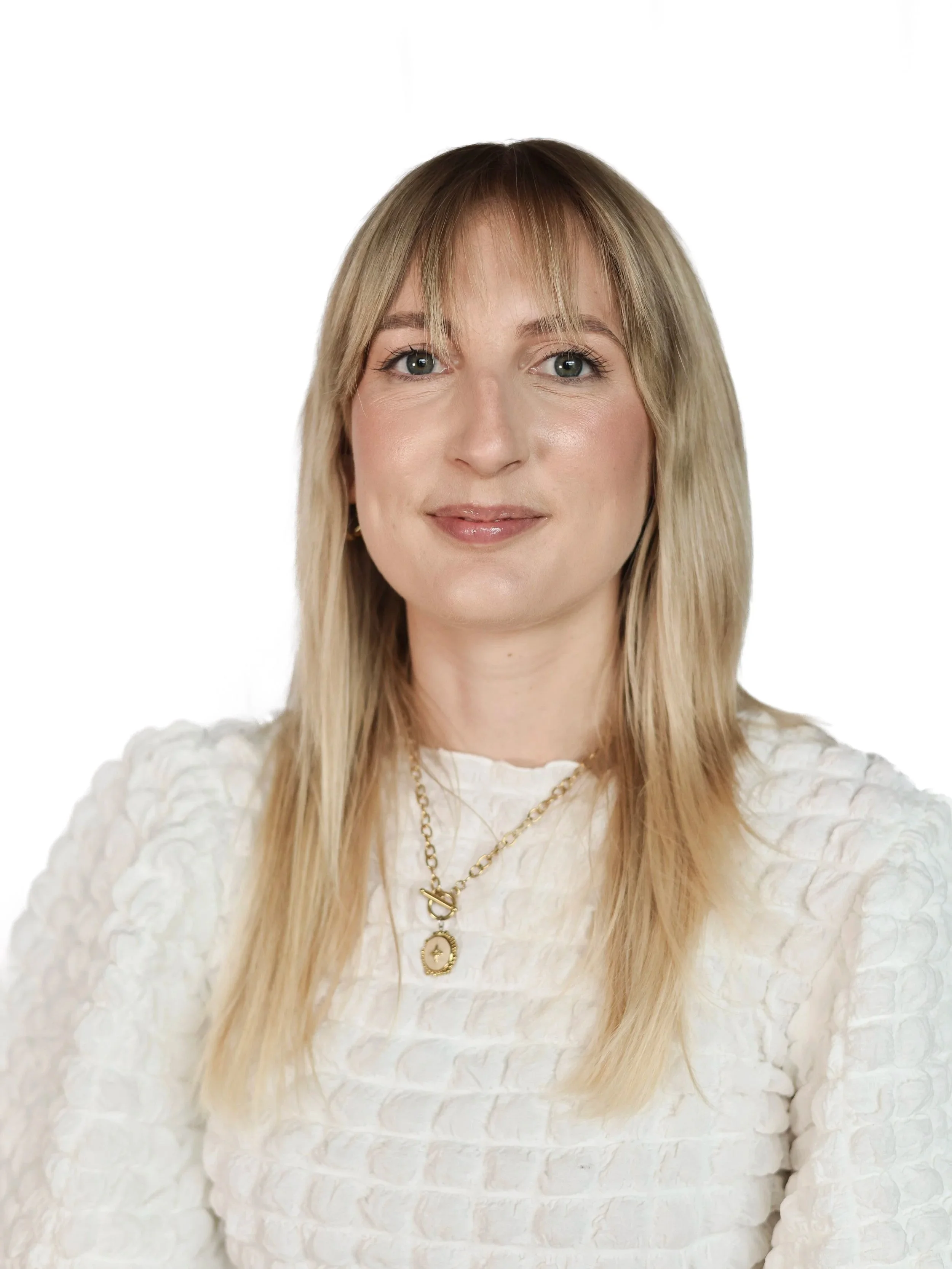 A woman with long blonde hair and blue eyes wearing a white textured top and layered gold necklaces, standing against a white background.