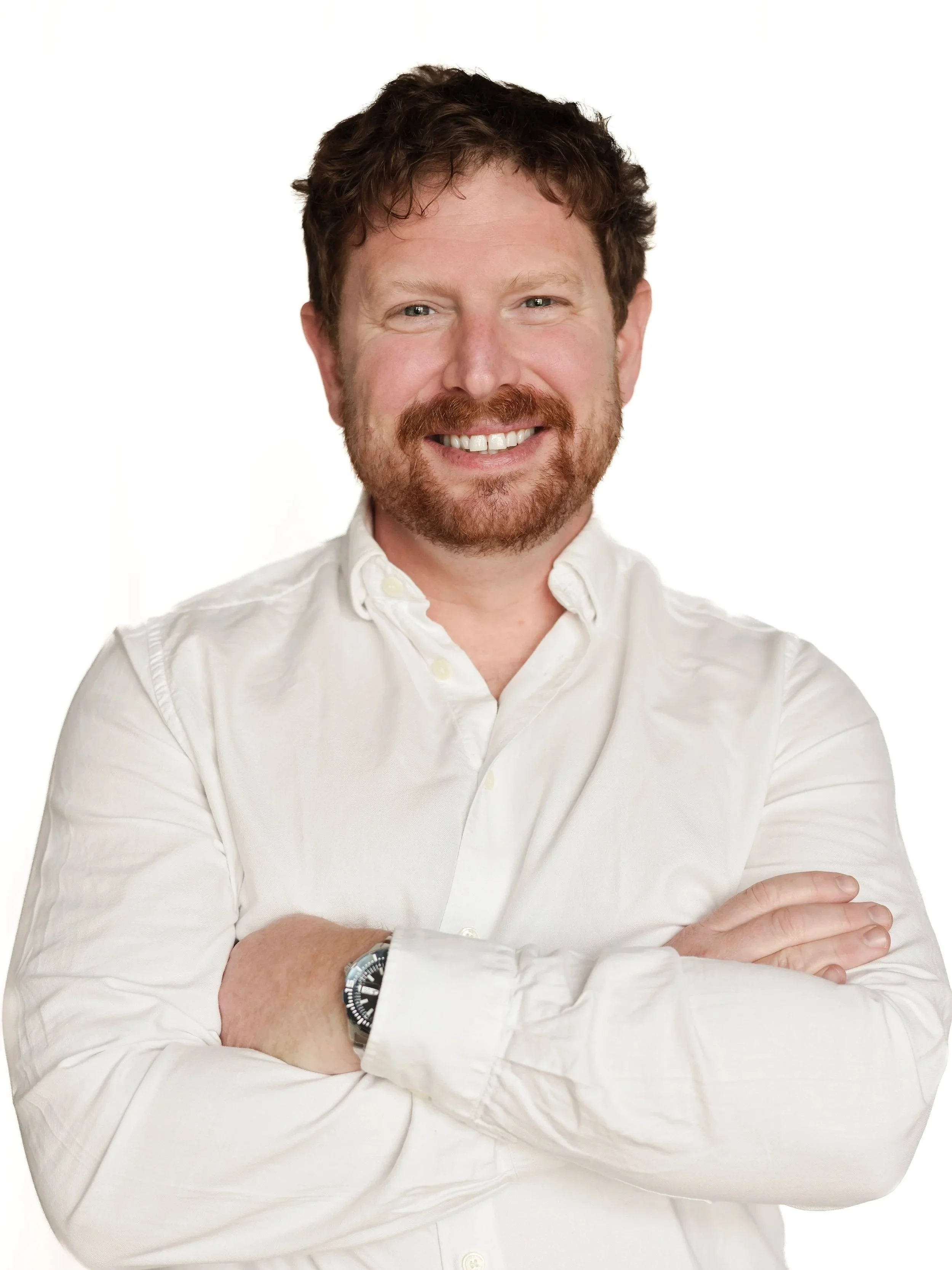 A man with red hair and a beard smiling, wearing a white button-up shirt and a watch, with arms crossed, against a plain white background.