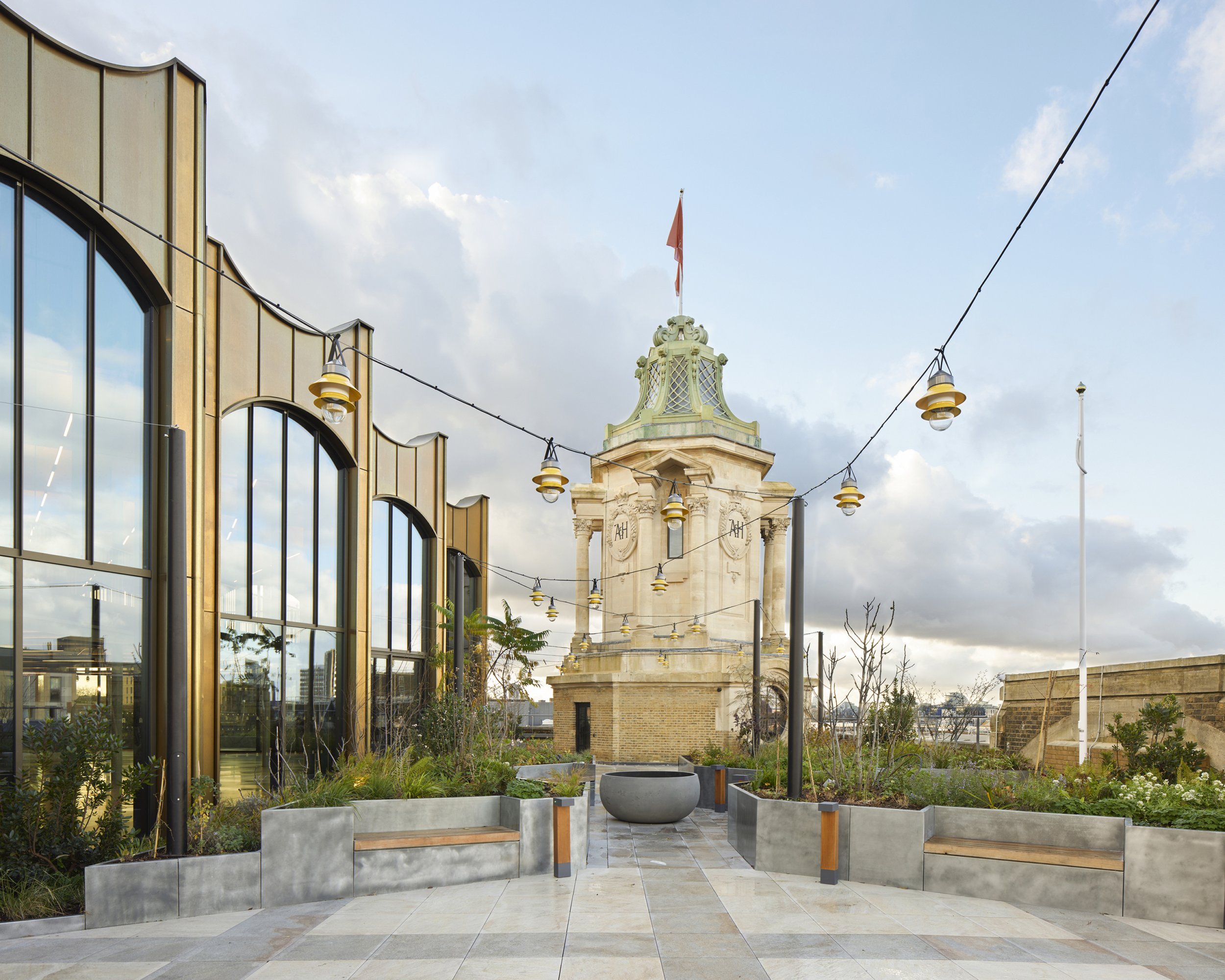 An urban outdoor rooftop garden with modern benches, planters, and string lights, with a historic building tower with a flag and decorative carvings in the background under a partly cloudy sky.