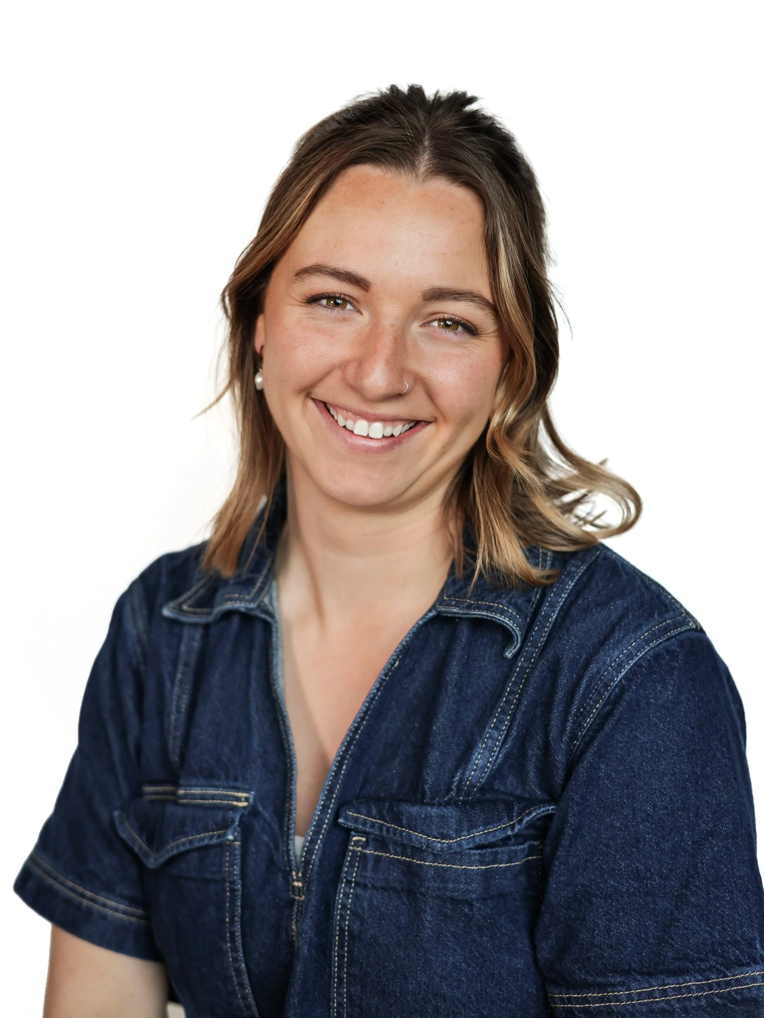 Smiling woman with shoulder-length wavy brown hair wearing a dark denim jacket against a plain white background.