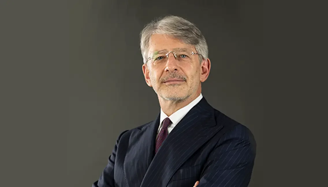 A middle-aged man with gray hair, glasses, and a beard, wearing a dark pinstripe suit and a white shirt, standing against a gray background.