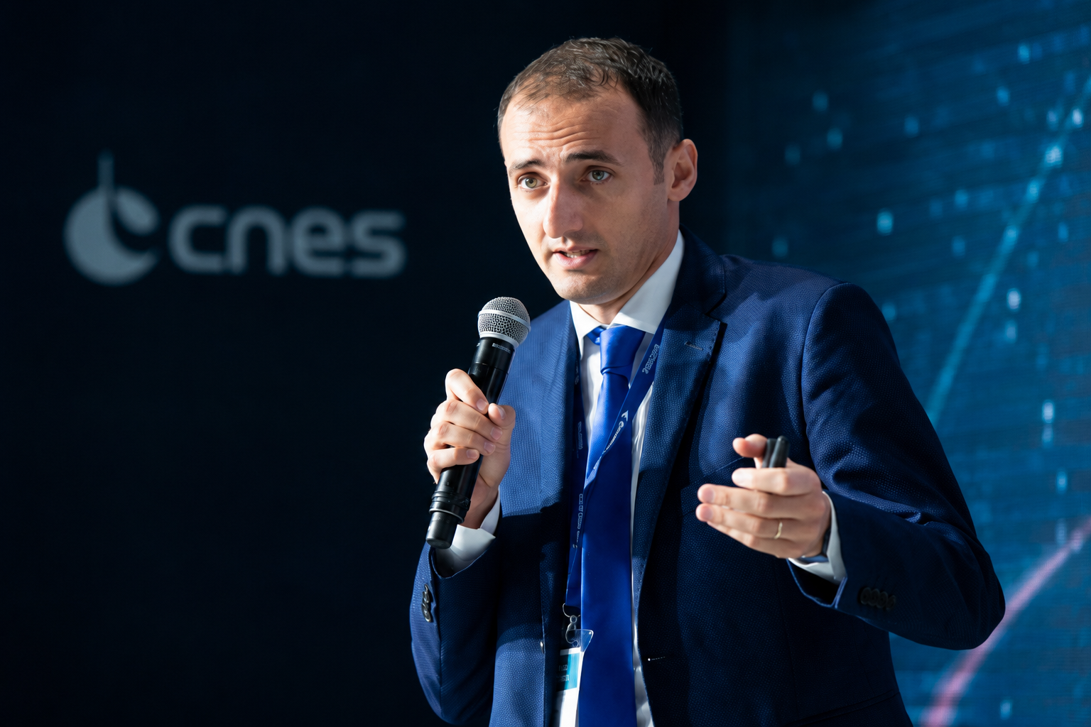 A man in a blue suit and tie holding a microphone and pointing during a presentation at a conference with a CNES logo in the background.
