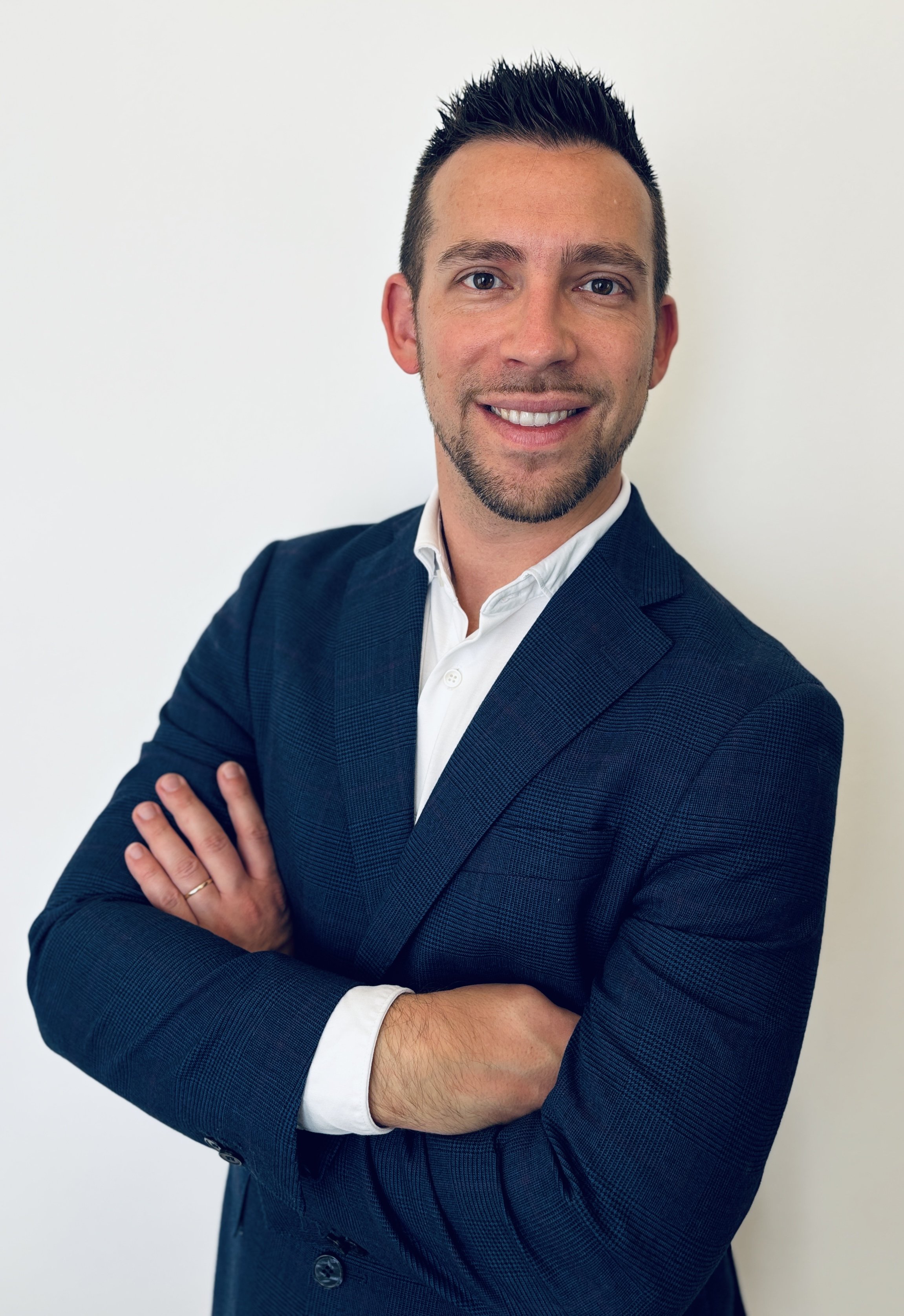 A young man with short dark hair, a goatee, and blue eyes, dressed in a navy blue blazer and white collared shirt, smiling with arms crossed against a plain white background.
