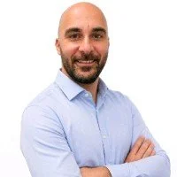 A man with a bald head and beard smiling, wearing a light blue button-up shirt, standing against a plain white background.