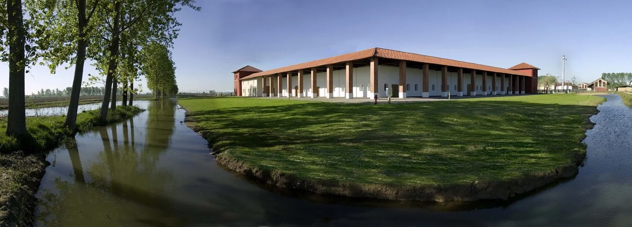 A large building with a red-tiled roof and multiple columns on a grassy lawn, surrounded by trees and water on both sides, under a clear blue sky.