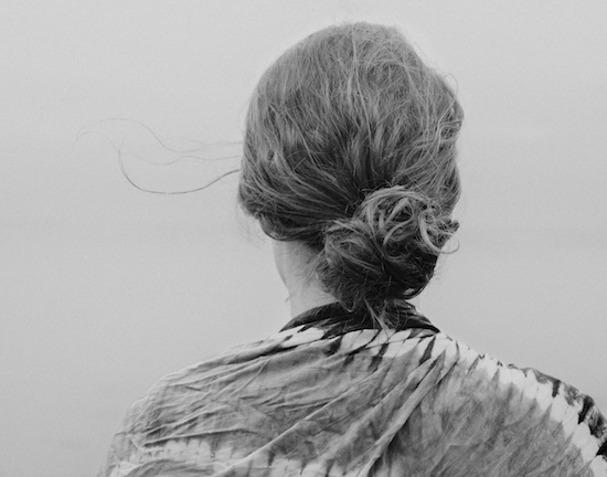 Back view of a woman with curly hair tied in a bun, wearing a patterned shawl, near a body of water in a black and white photograph.