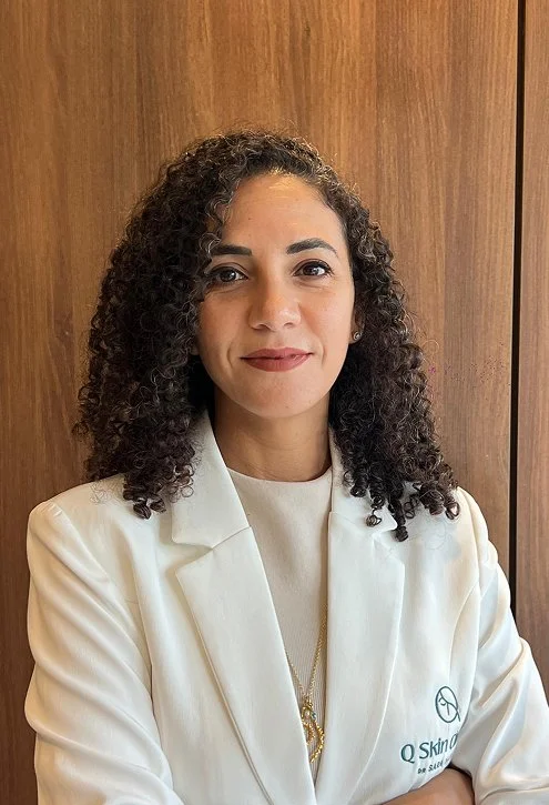 Professional woman with curly hair in a white blazer standing in front of a wooden wall.