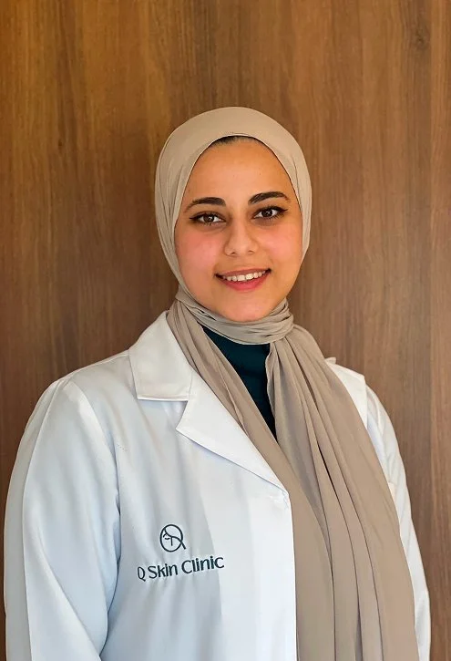 A woman wearing a beige hijab and white lab coat with a logo and text that reads "Q Skin Clinic" stands in front of a wooden background.