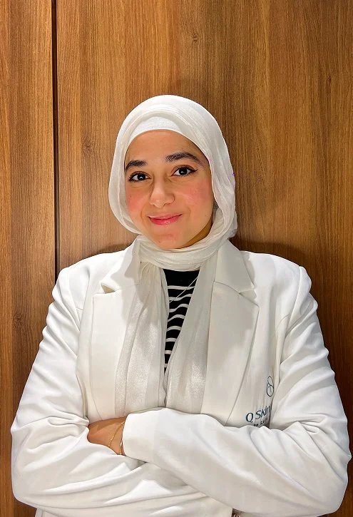 Woman with a white headscarf and white coat, standing against a wooden background, smiling with arms crossed.