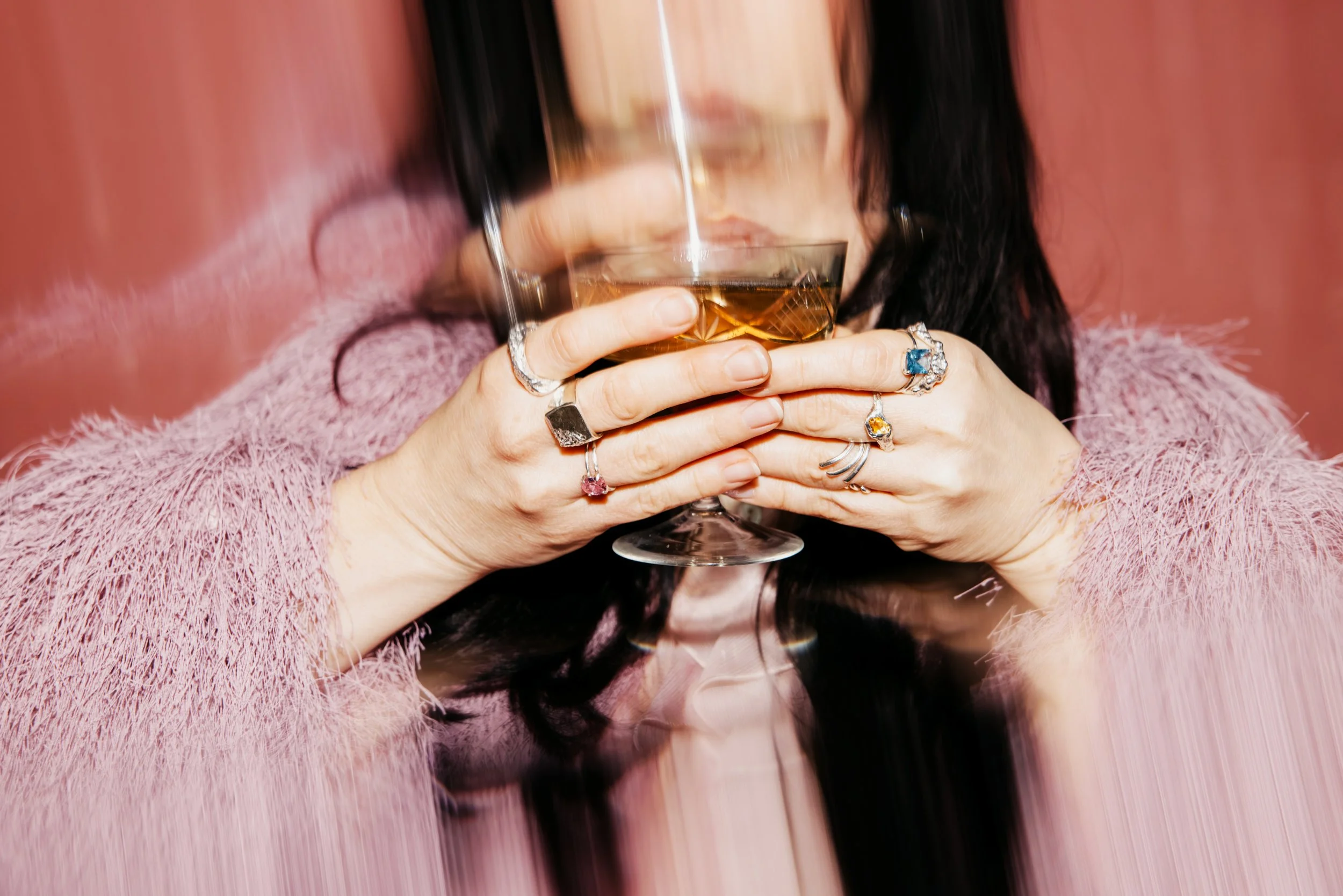 Eine Frau mit dunklen Haaren trägt mehrere Ringe und hält ein Weinglas mit einem goldfarbenen Getränk in beiden Händen. Sie trägt einen pinken, flauschigen Pullover.