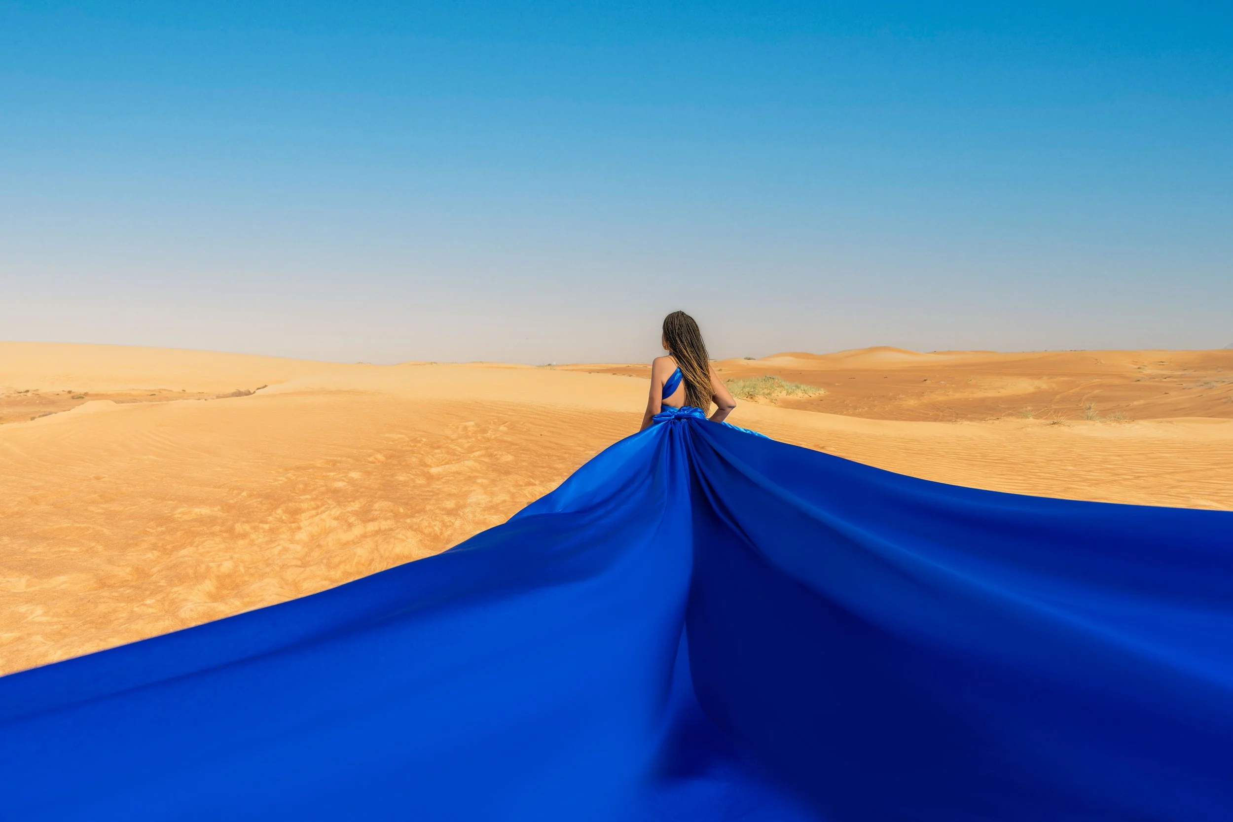 Woman in a flowing blue dress standing in a desert landscape under a clear blue sky.